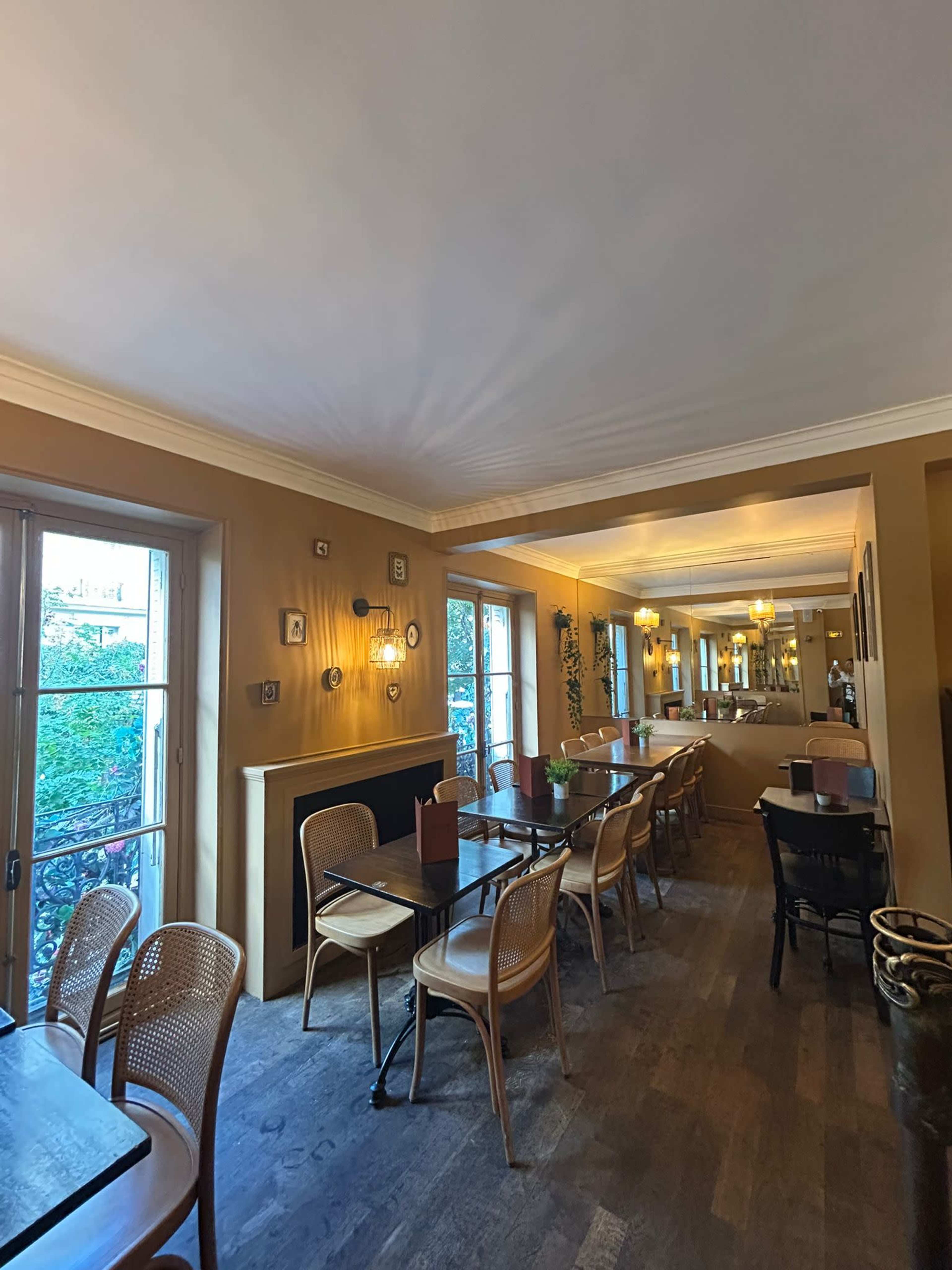 The image shows a restaurant interior with wooden floors, tables with chairs arranged in rows, and large windows allowing natural light.