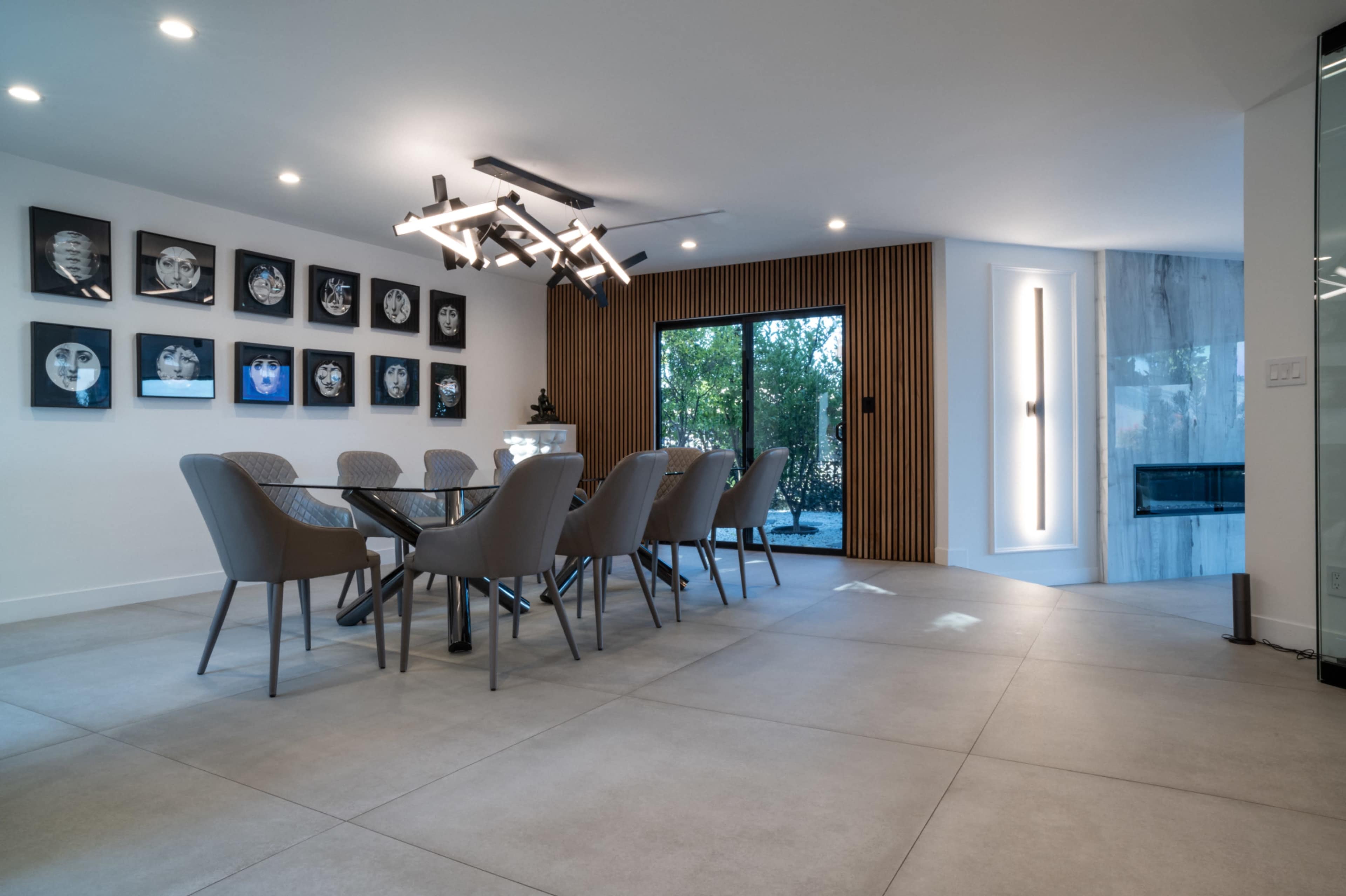 A modern dining area features a glass table surrounded by gray chairs, with a gallery wall of framed artwork and large windows opening to an outdoor space.