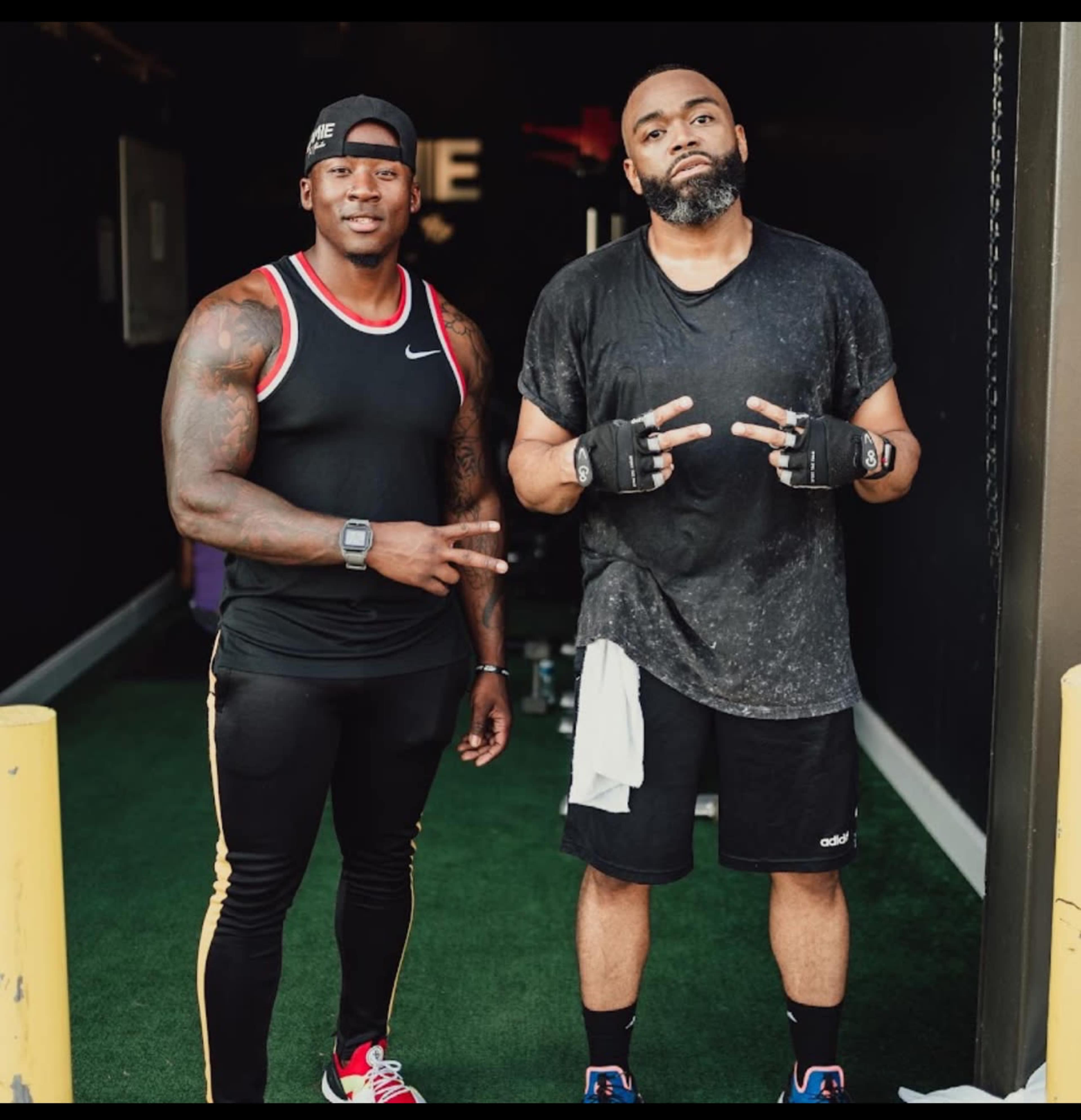 Two men stand in a gym entrance, one in athletic wear and the other in a tank top and shorts, both displaying peace signs.