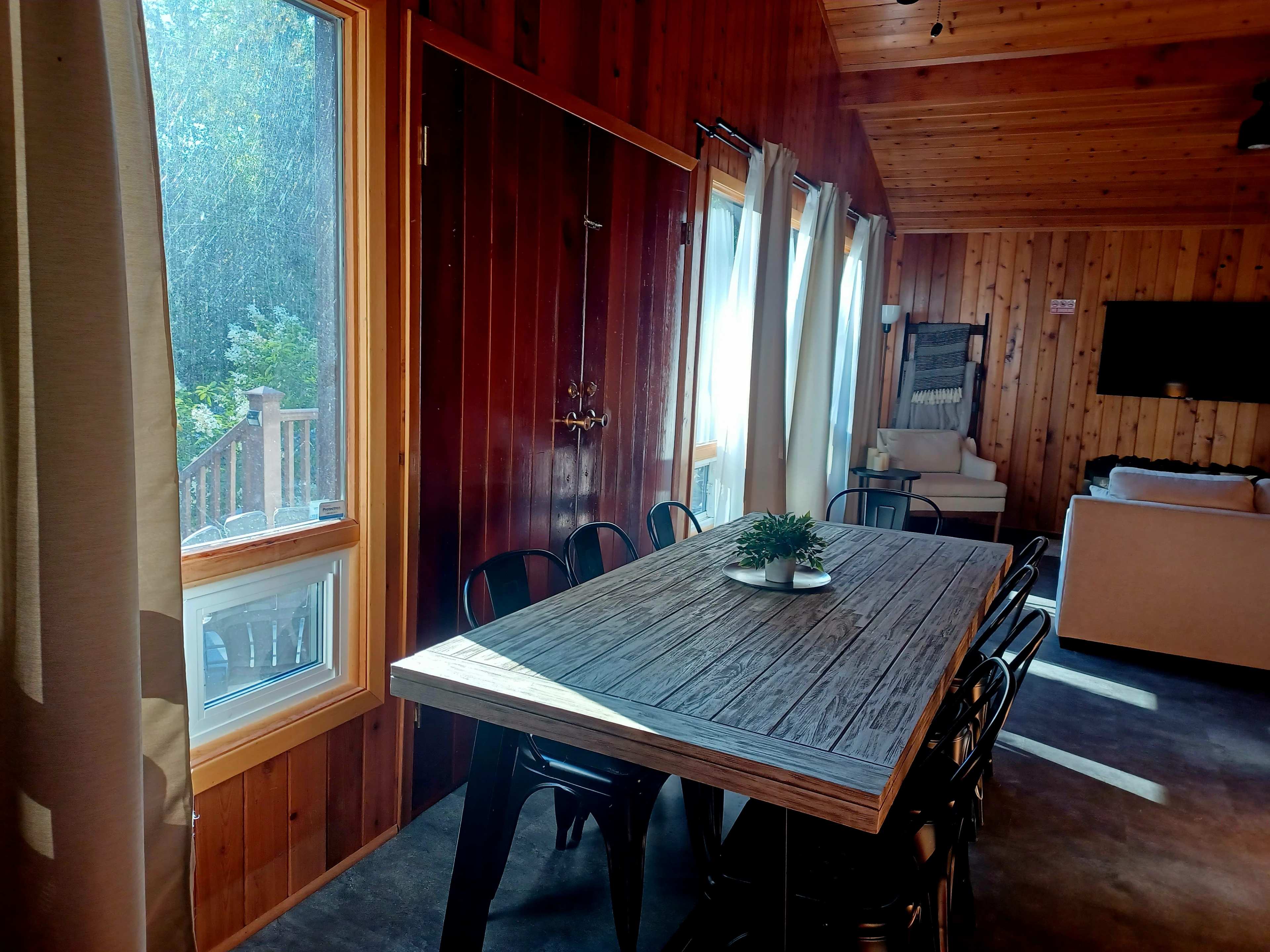 A large wooden dining table is set in a well-lit room featuring wooden walls and a view of the outdoors through a window.