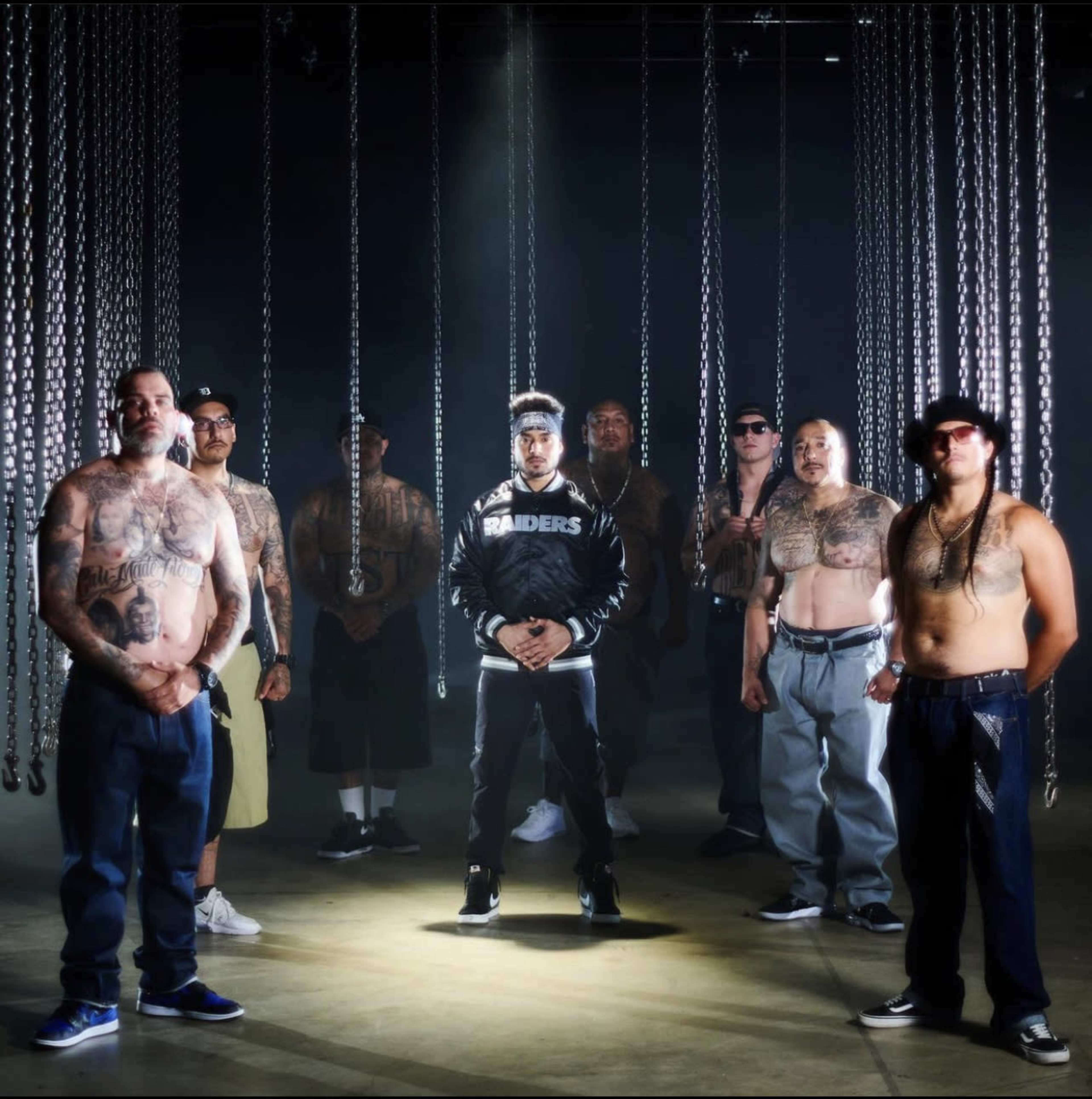 A group of eight men stands in a dimly lit space with chains hanging from the ceiling, showcasing various tattoos and dressed in casual clothing.