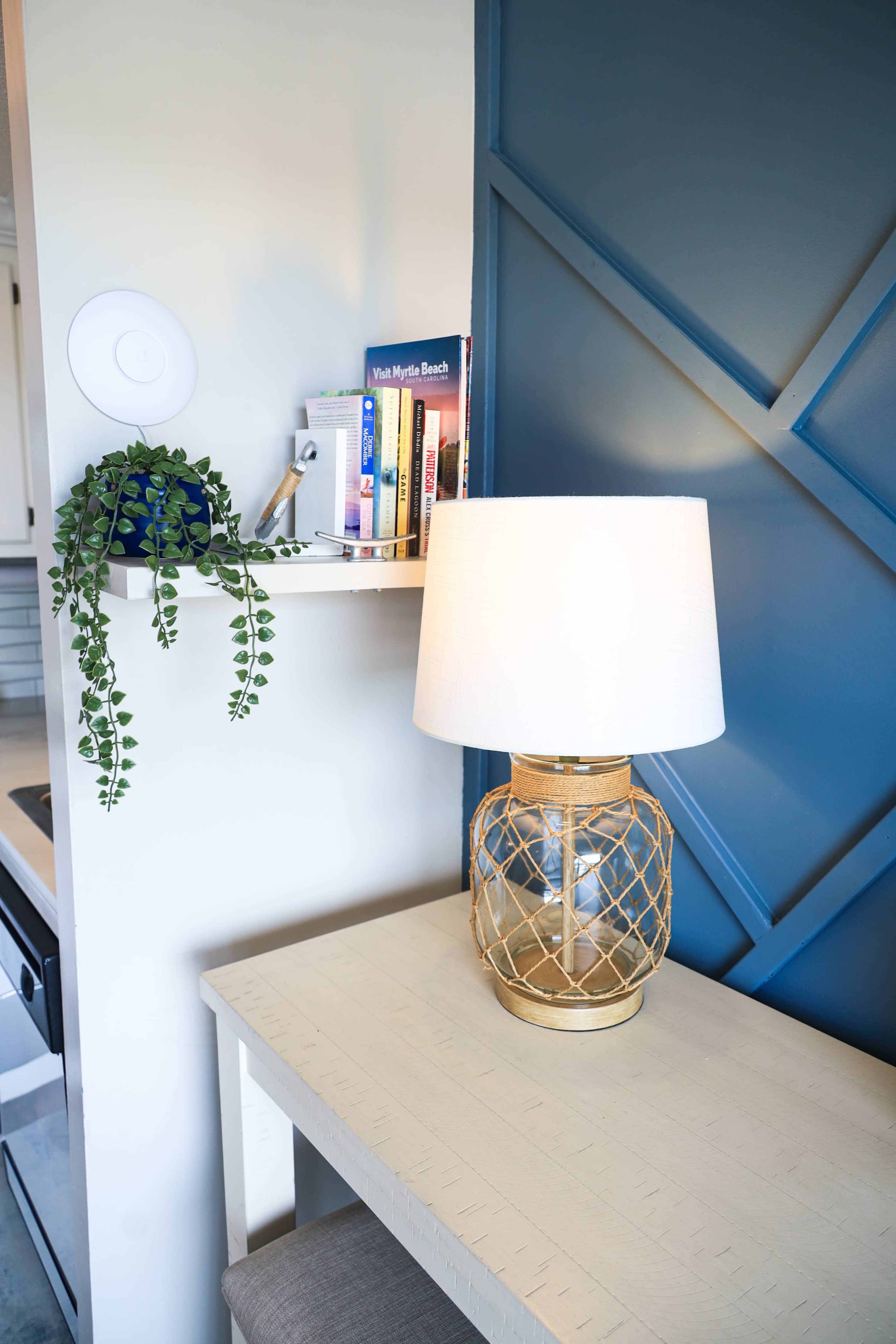 A table with a gold lamp and a shelf holding books and a potted plant is positioned against a blue wall with a geometric design.