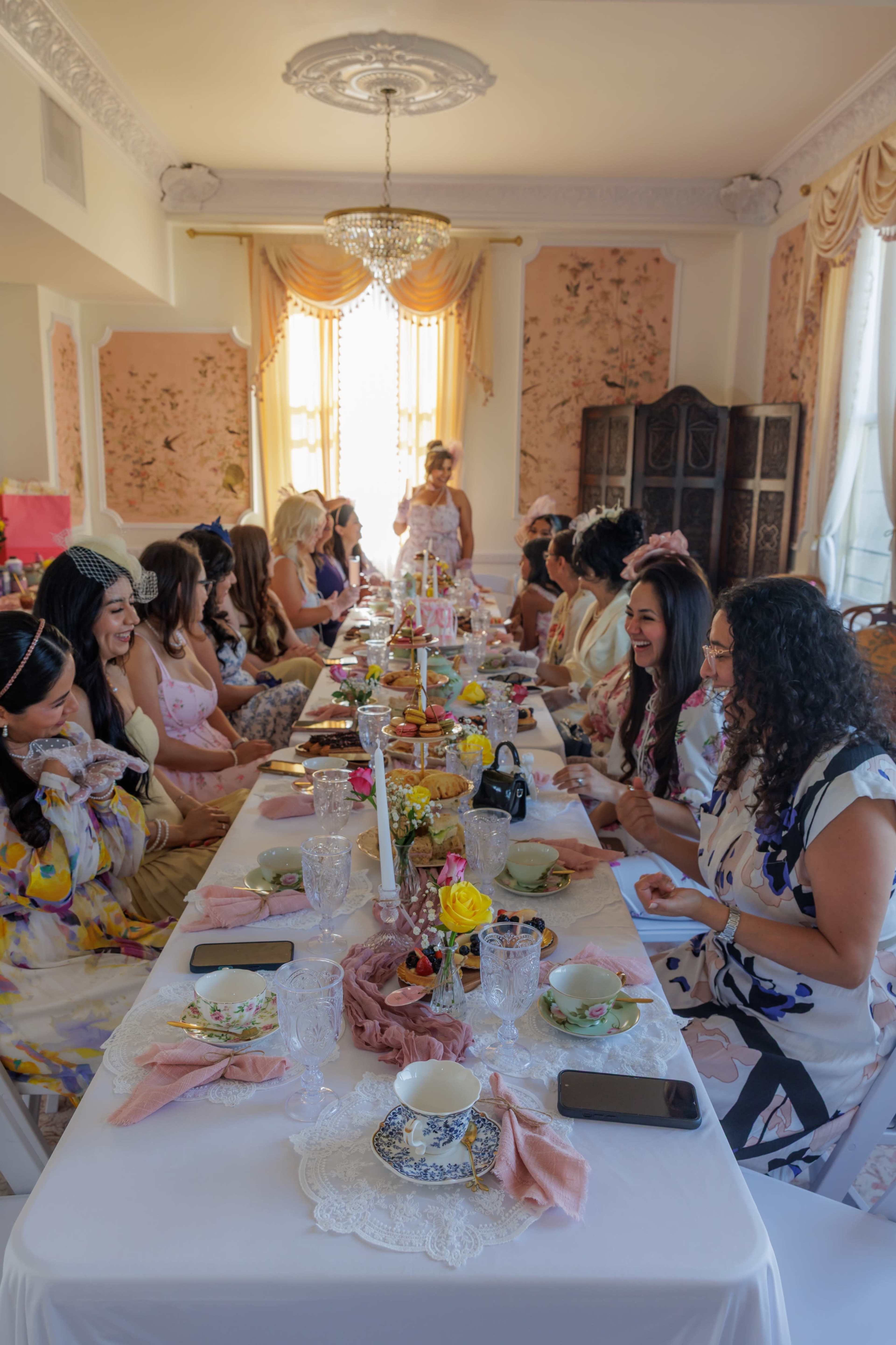 A long table is set in a beautifully decorated room, filled with women enjoying a tea party with a variety of food and drink.