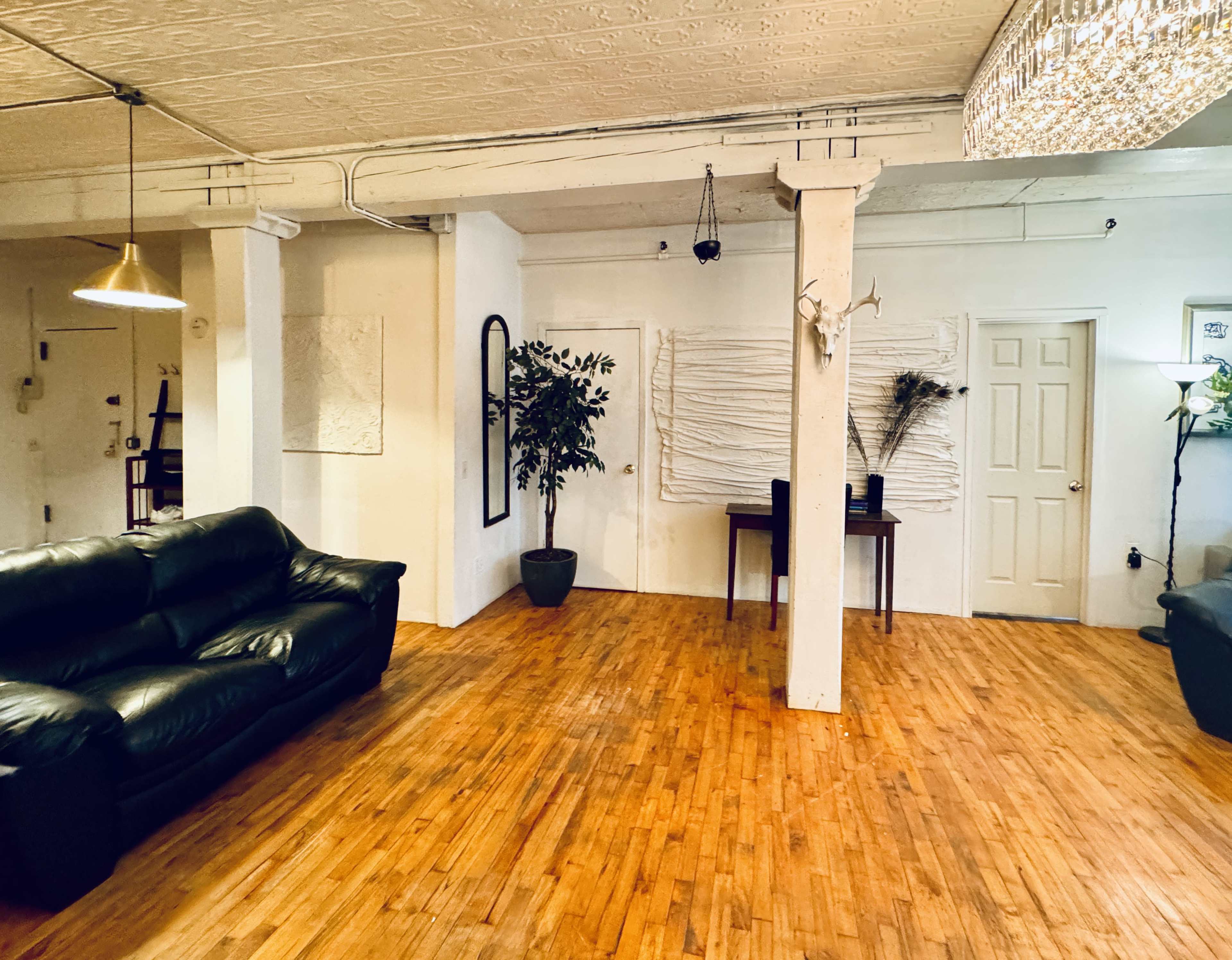 The image shows a spacious, minimalist living area with wooden flooring, a black leather sofa, a small dining table, and decorative wall textures.