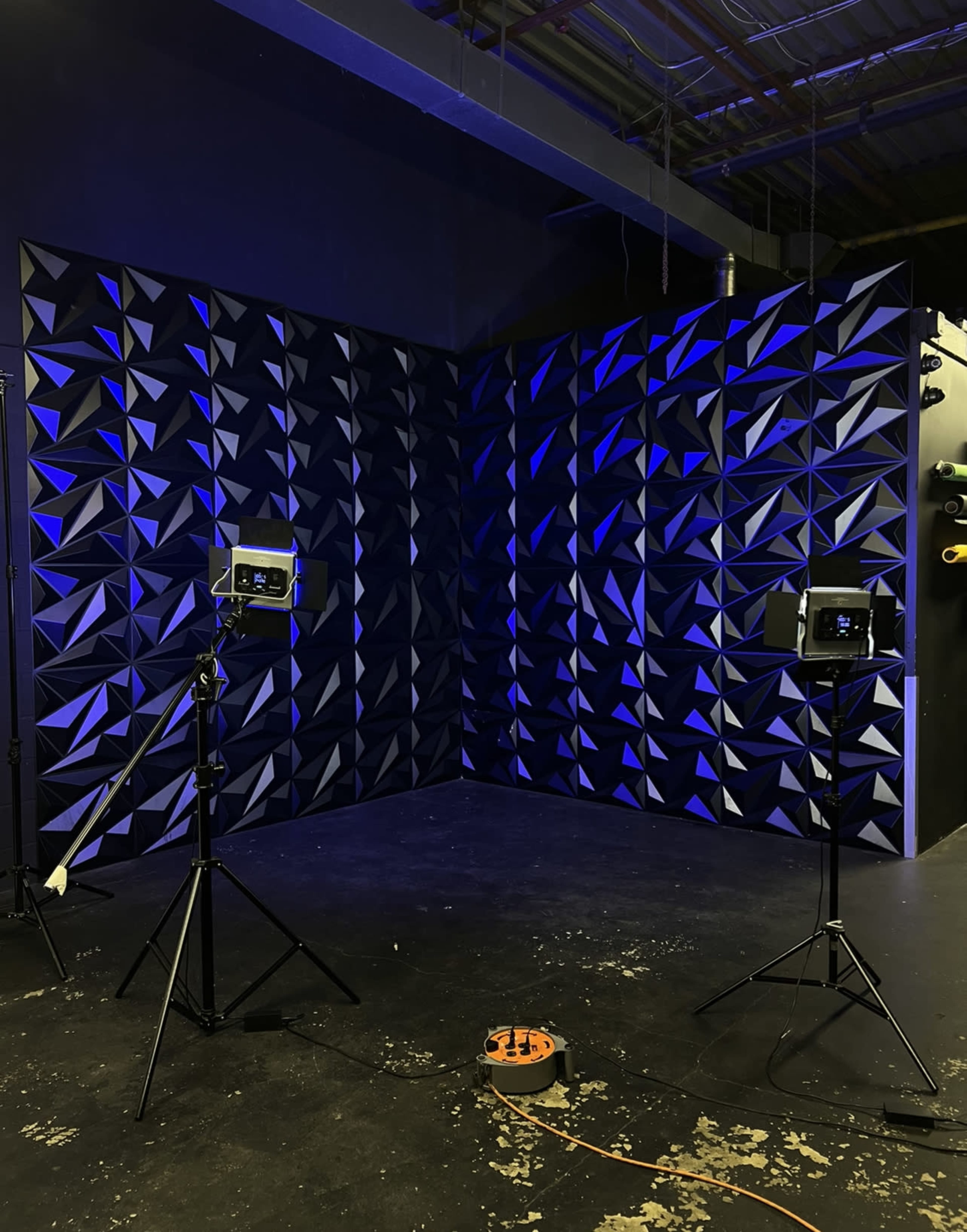 The image shows a dark room with a textured, geometric wall and two lights on tripods illuminating the corner.