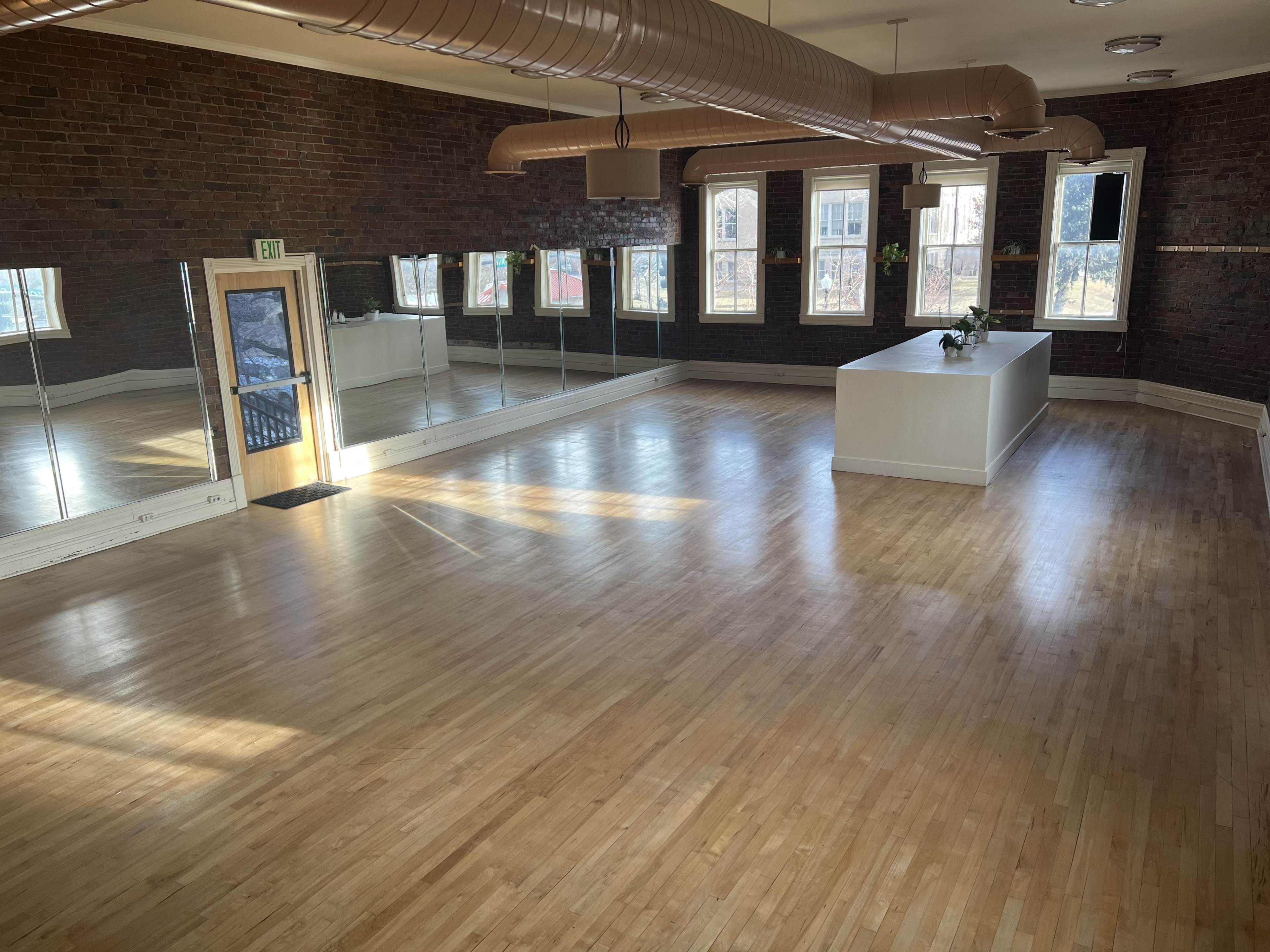 The image shows a spacious, empty room with wooden floors, large windows, and a central white pedestal, surrounded by exposed brick walls and reflections in a mirror.