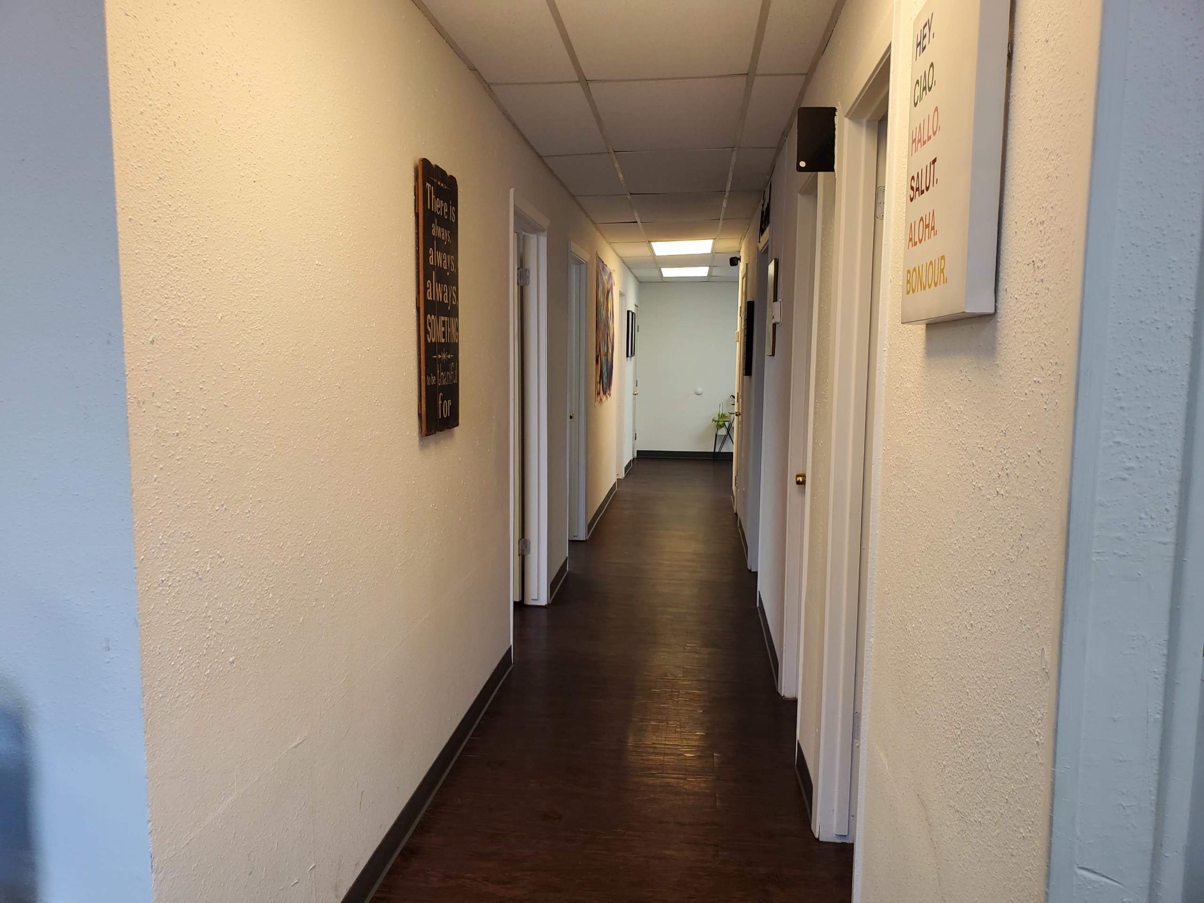 The image shows a long hallway with white walls, wooden flooring, and doors lining both sides, along with framed signs on the walls.