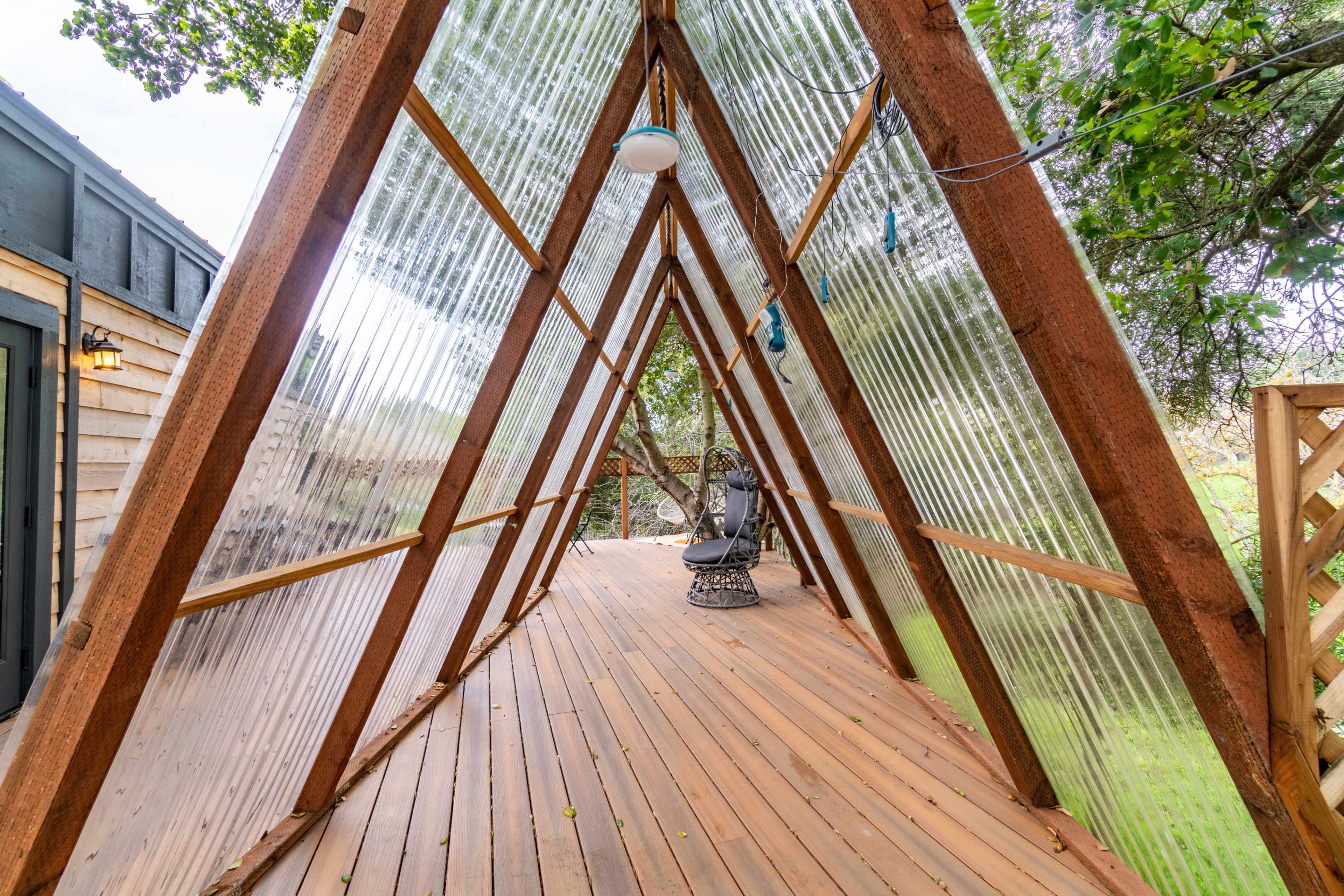 The image shows a wooden deck in an A-frame structure with transparent walls, surrounded by greenery.
