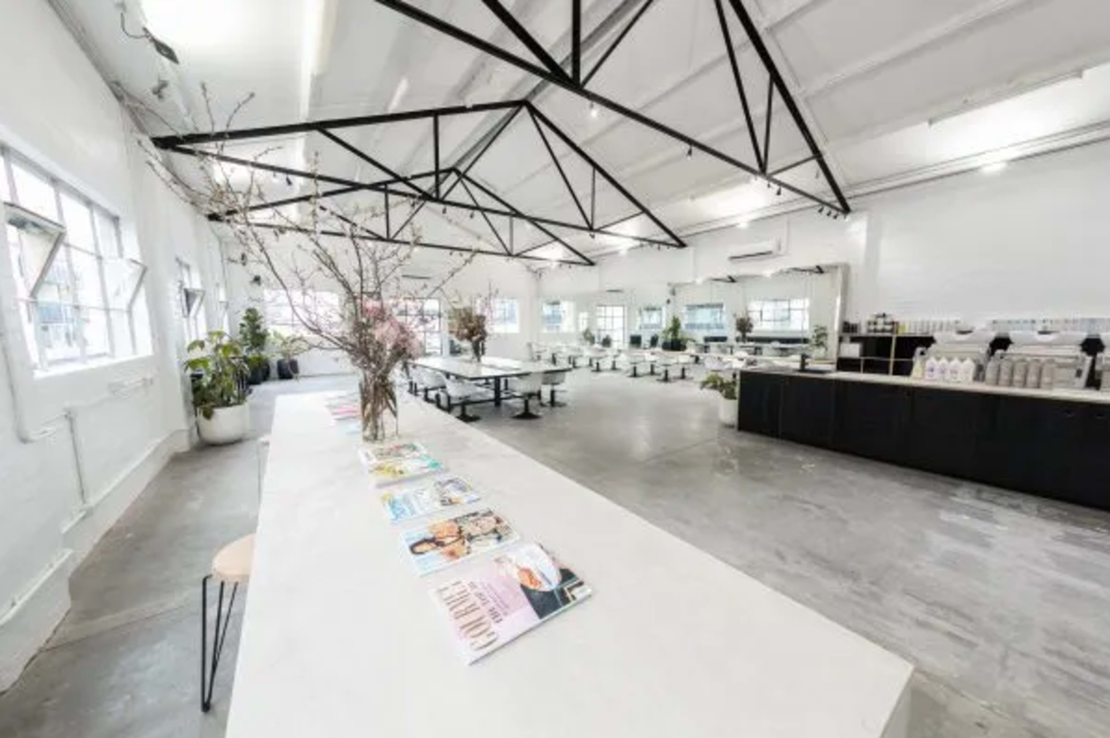 The image depicts a spacious, modern interior with a long white table, black seating, and large windows, accented by minimalistic decor and magazines displayed along the tabletop.
