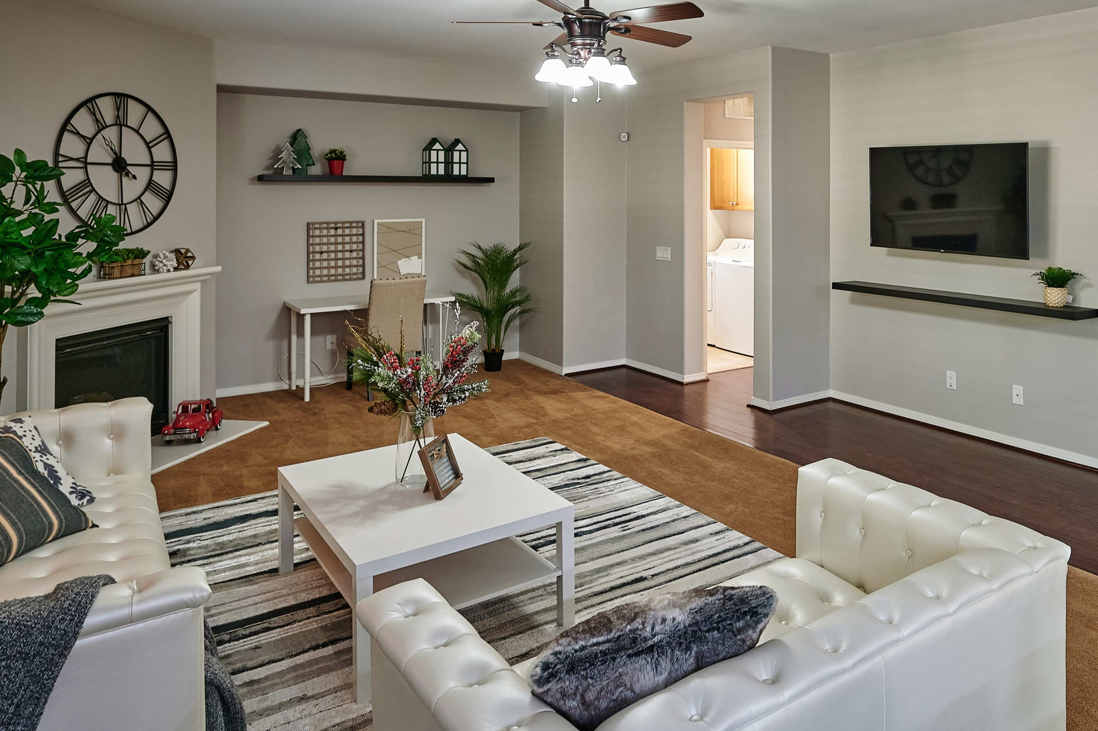 The living room features two white sofas facing a coffee table, with a clock on the wall, a fireplace, and a small desk in the background.