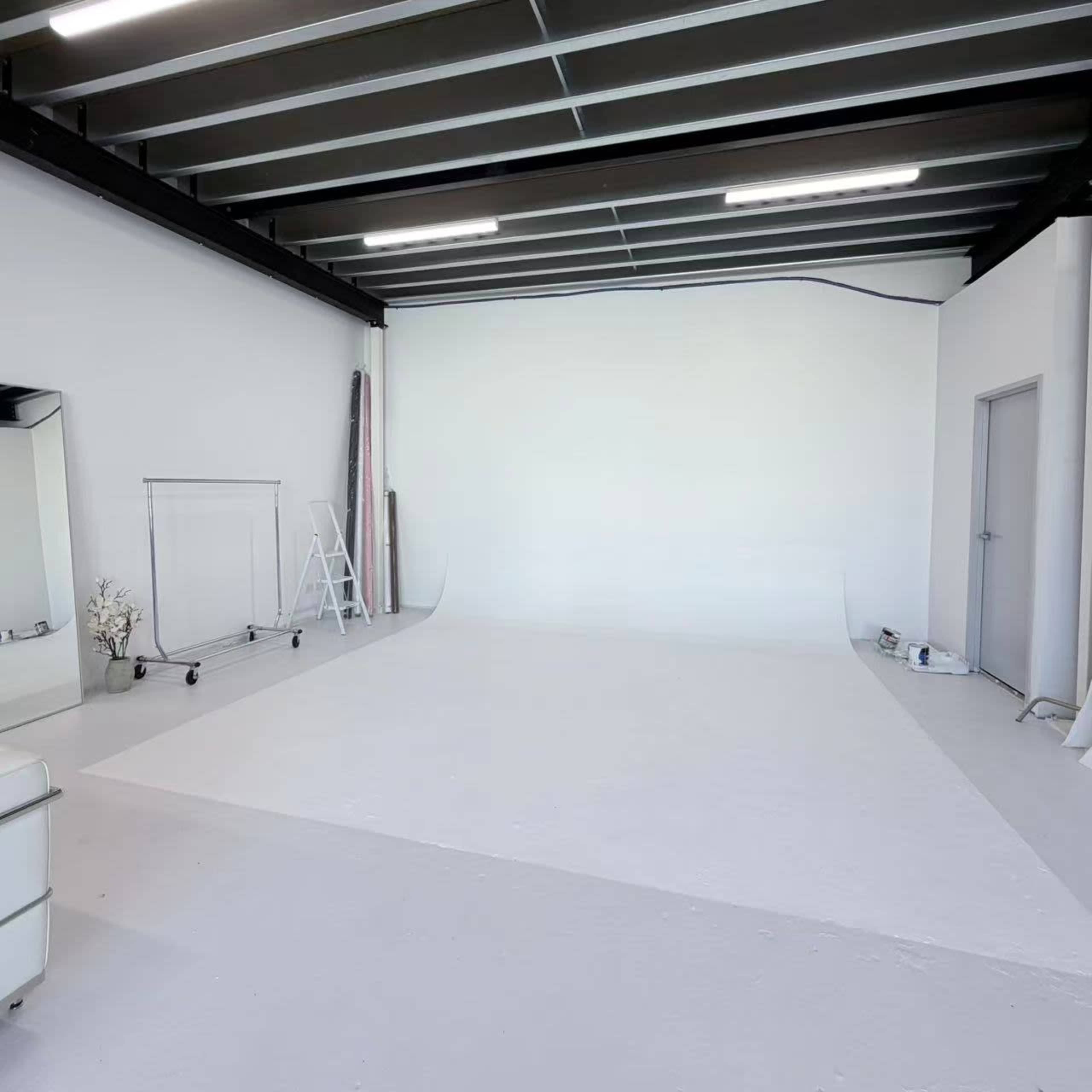 The image shows a spacious, minimalistic studio with a white backdrop, a folding ladder, and a mirror against one wall.