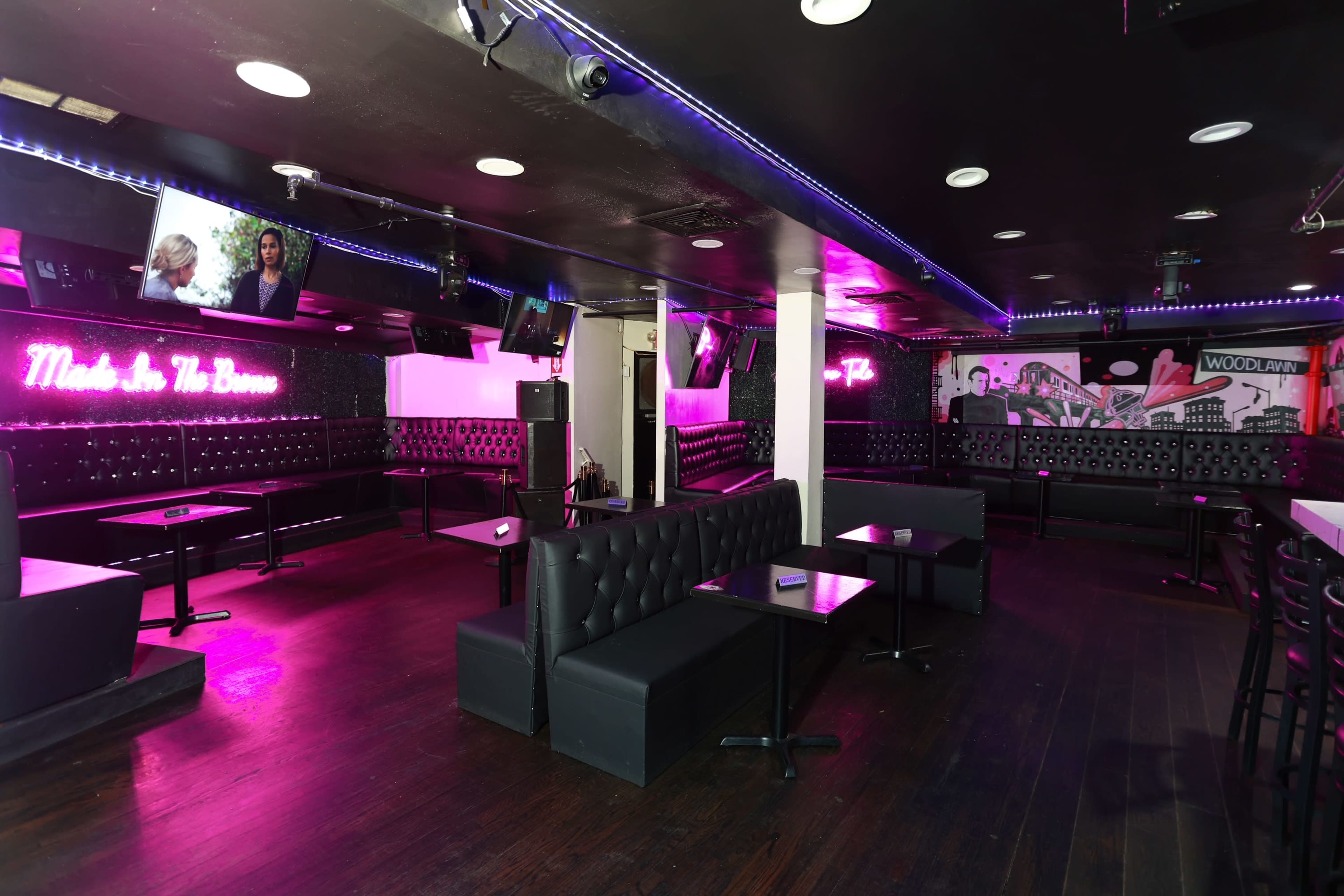 The image shows a dimly lit lounge area with black seating, purple neon lighting, and several television screens on the walls.