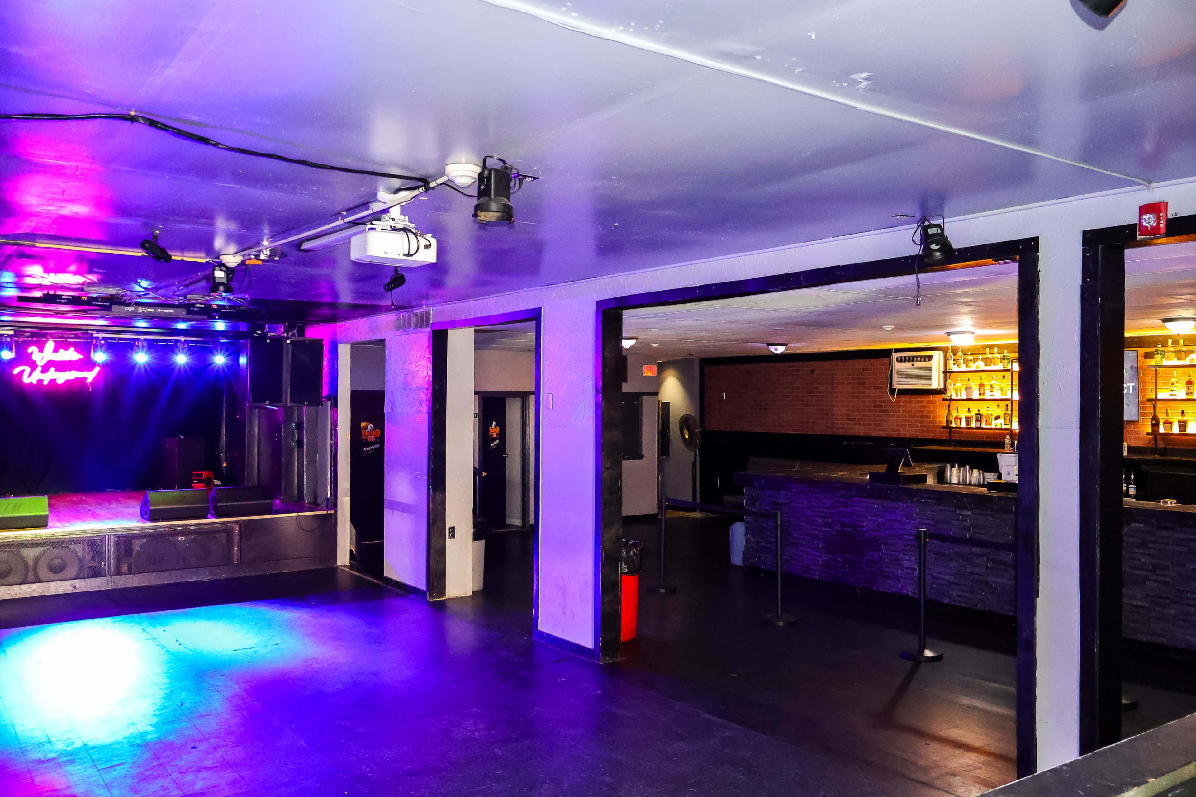 An empty nightclub interior featuring a stage with colorful lights and a bar area in the background.