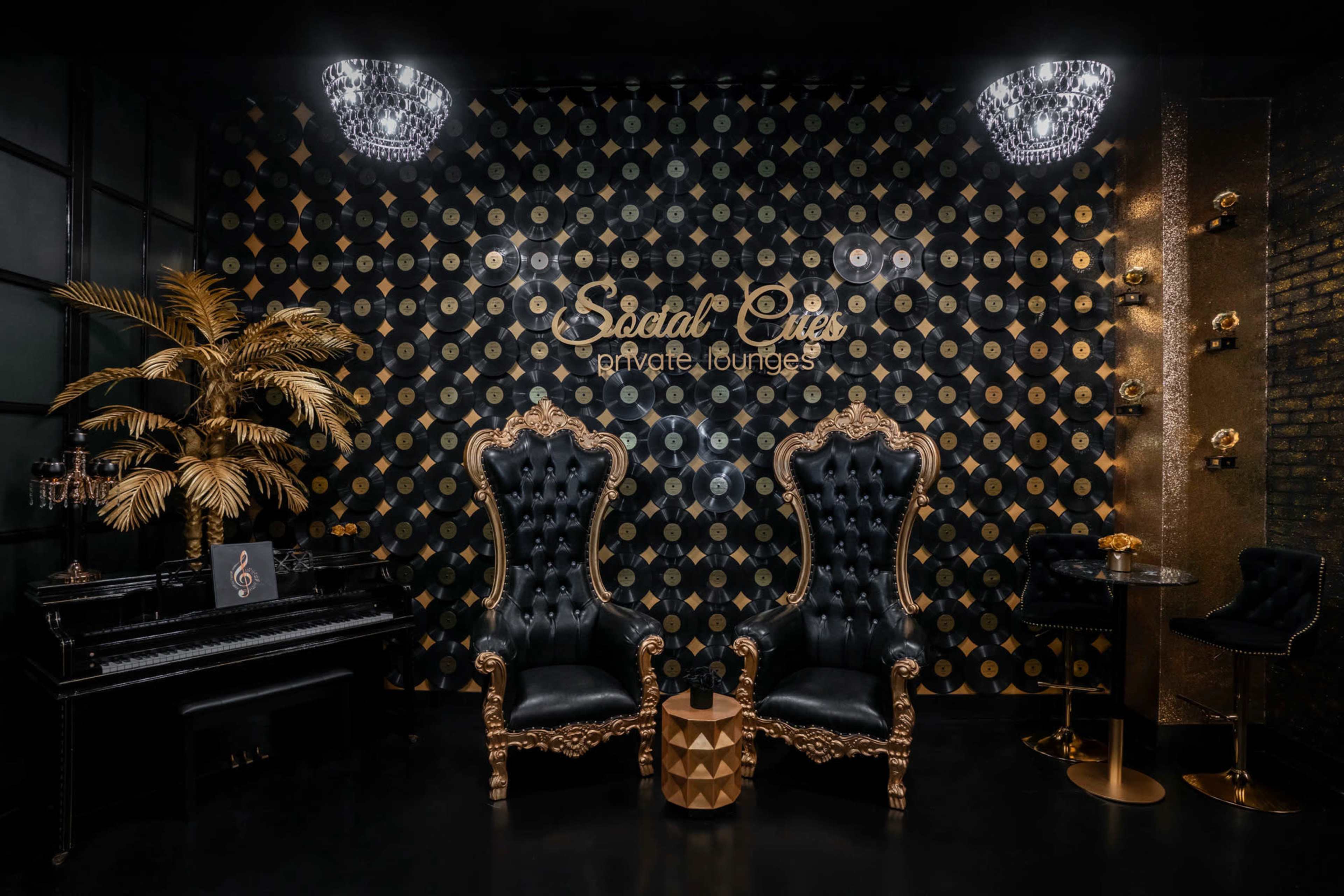 The image shows an elegant lounge area featuring two ornate black chairs, a piano, and a wall decorated with gold patterns and a logo.