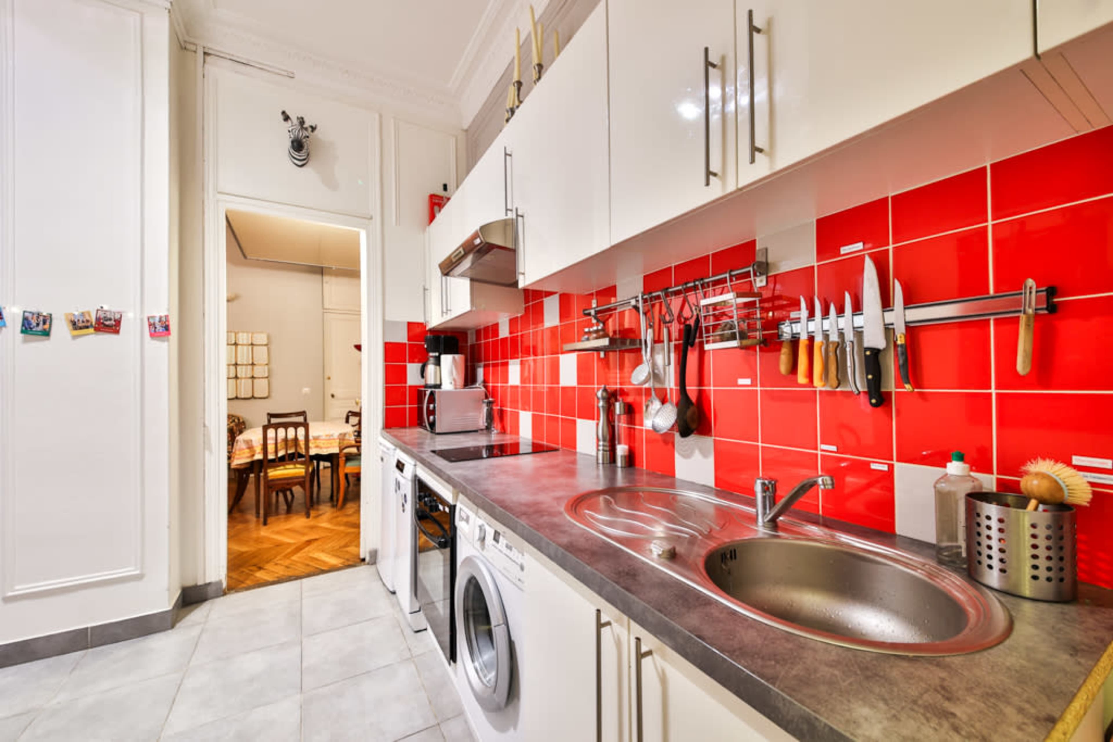 The image shows a modern kitchen with red tiled walls, stainless steel appliances, and a view into a dining area through an open doorway.
