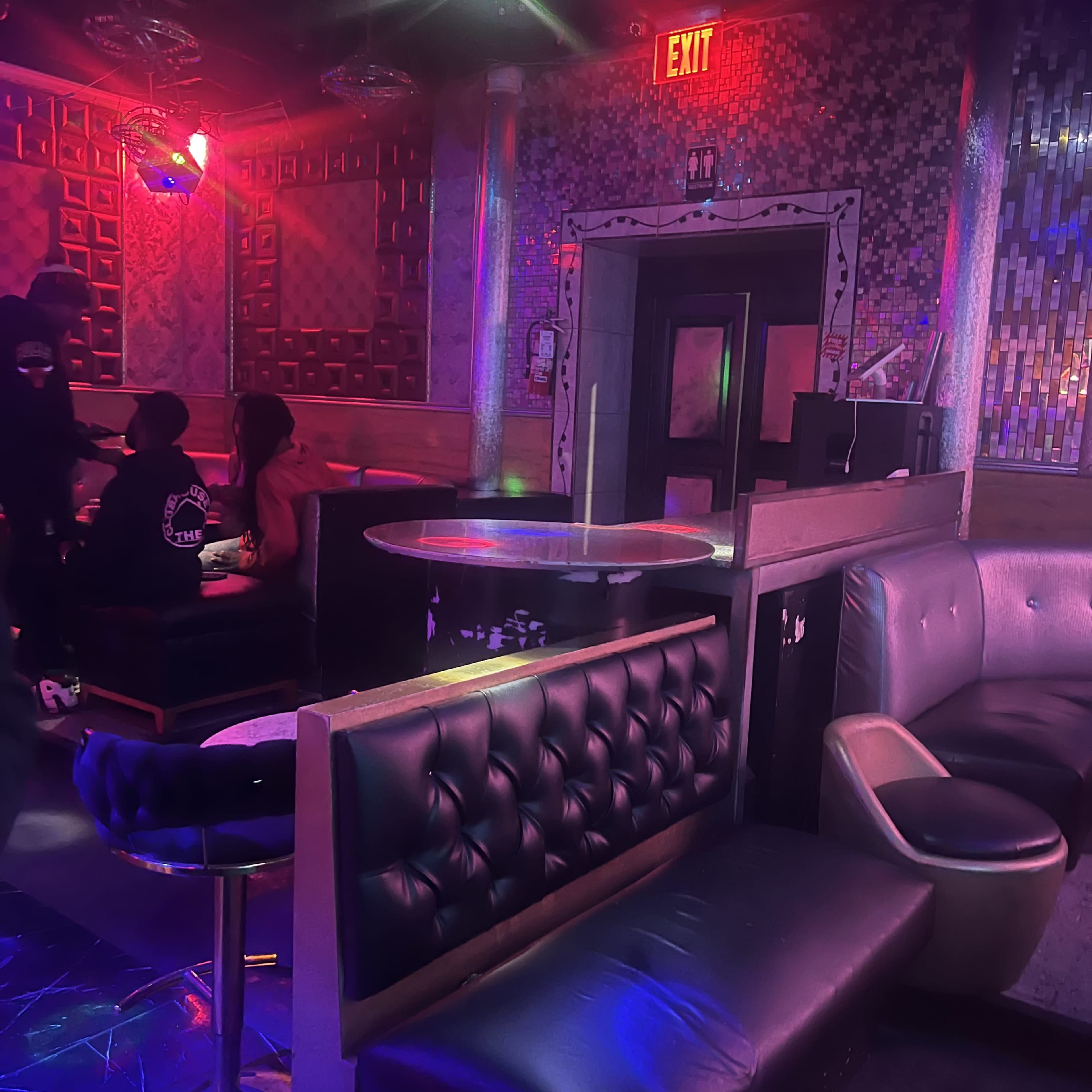 The image shows a dimly lit lounge area with upholstered seating, a small table, and colorful lighting effects.