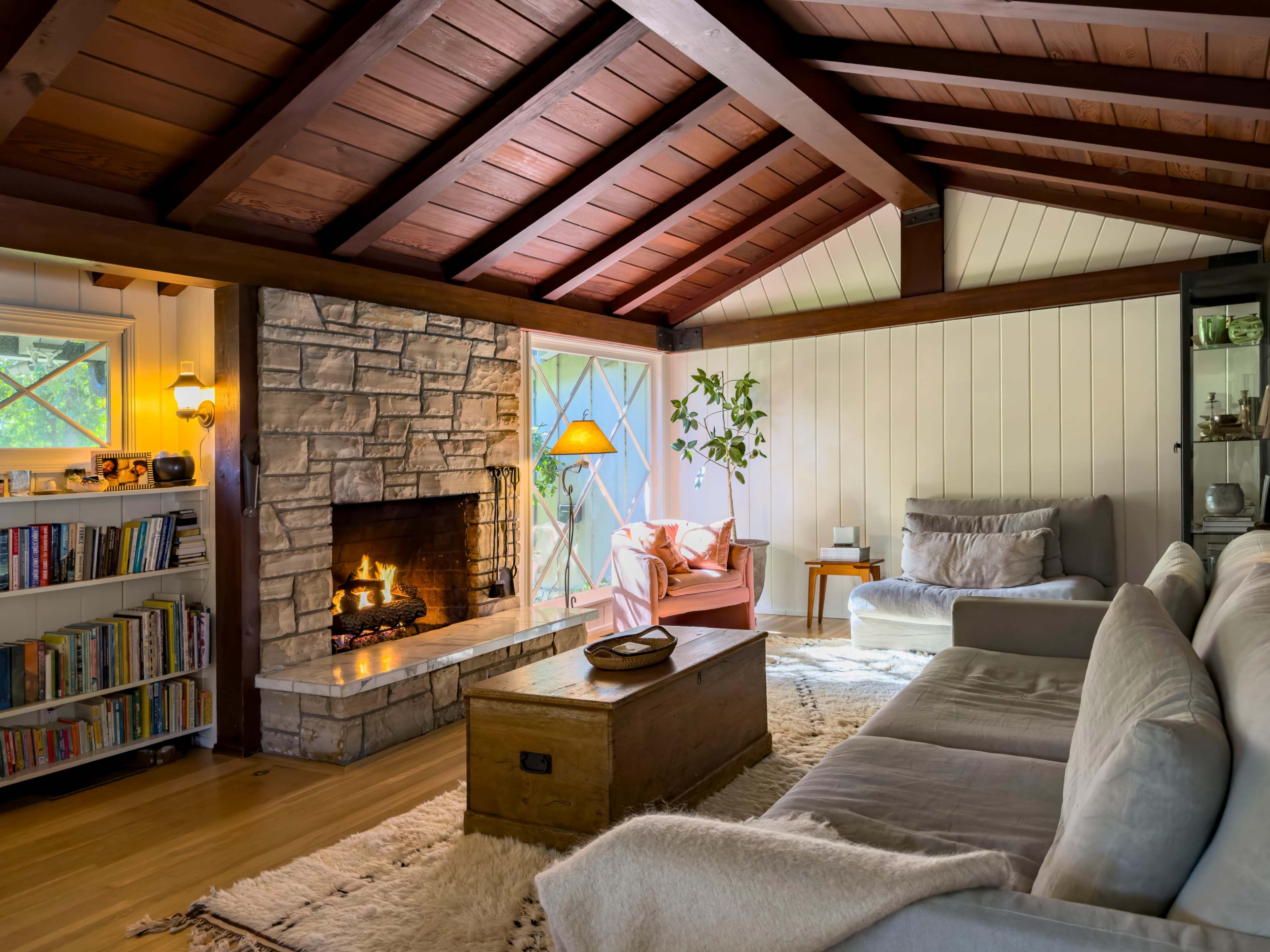 The living room features a stone fireplace, wooden beam ceiling, and cozy seating arrangements next to large windows.