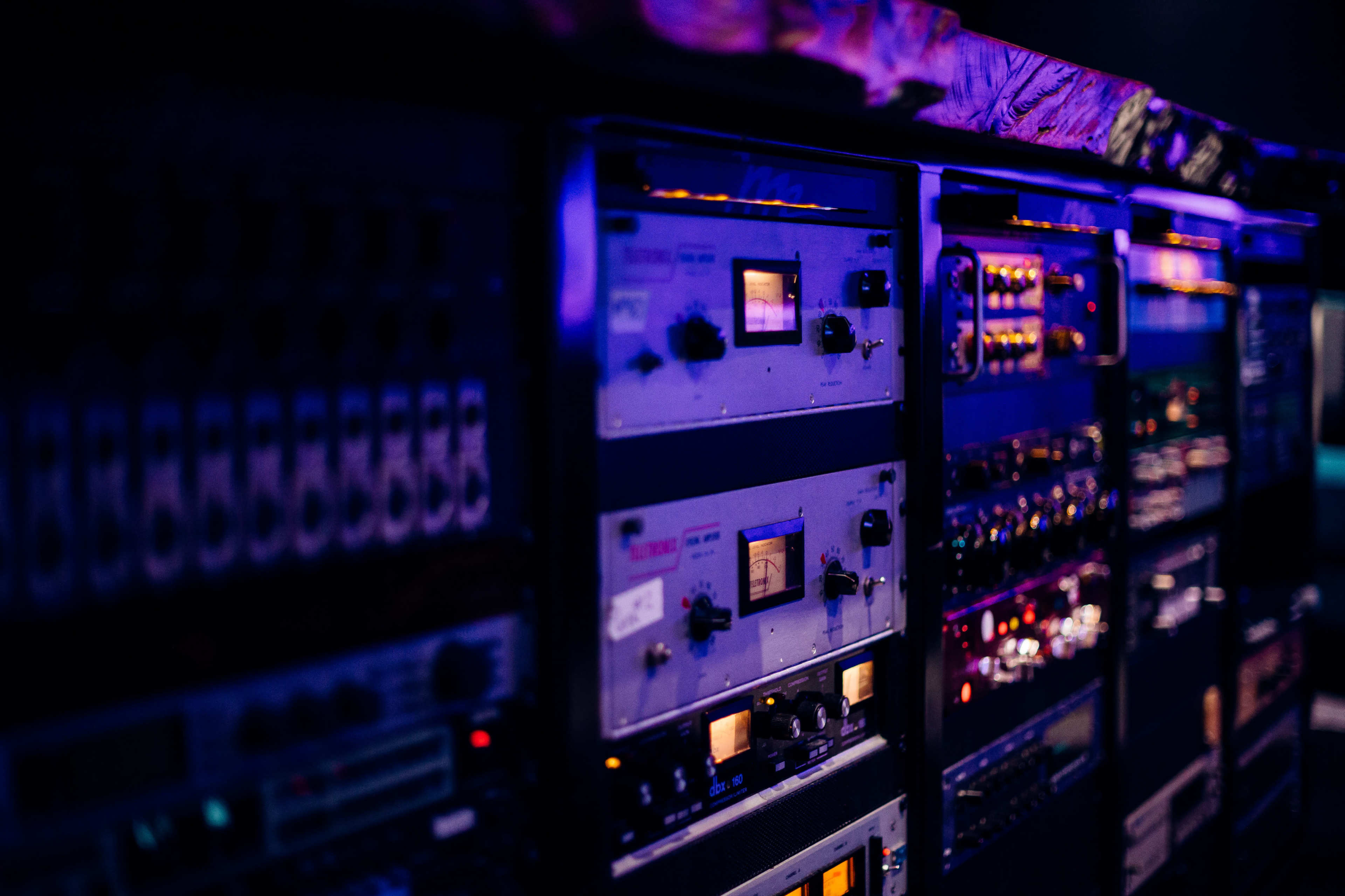 A row of audio equipment and sound mixers illuminated by dim lighting.