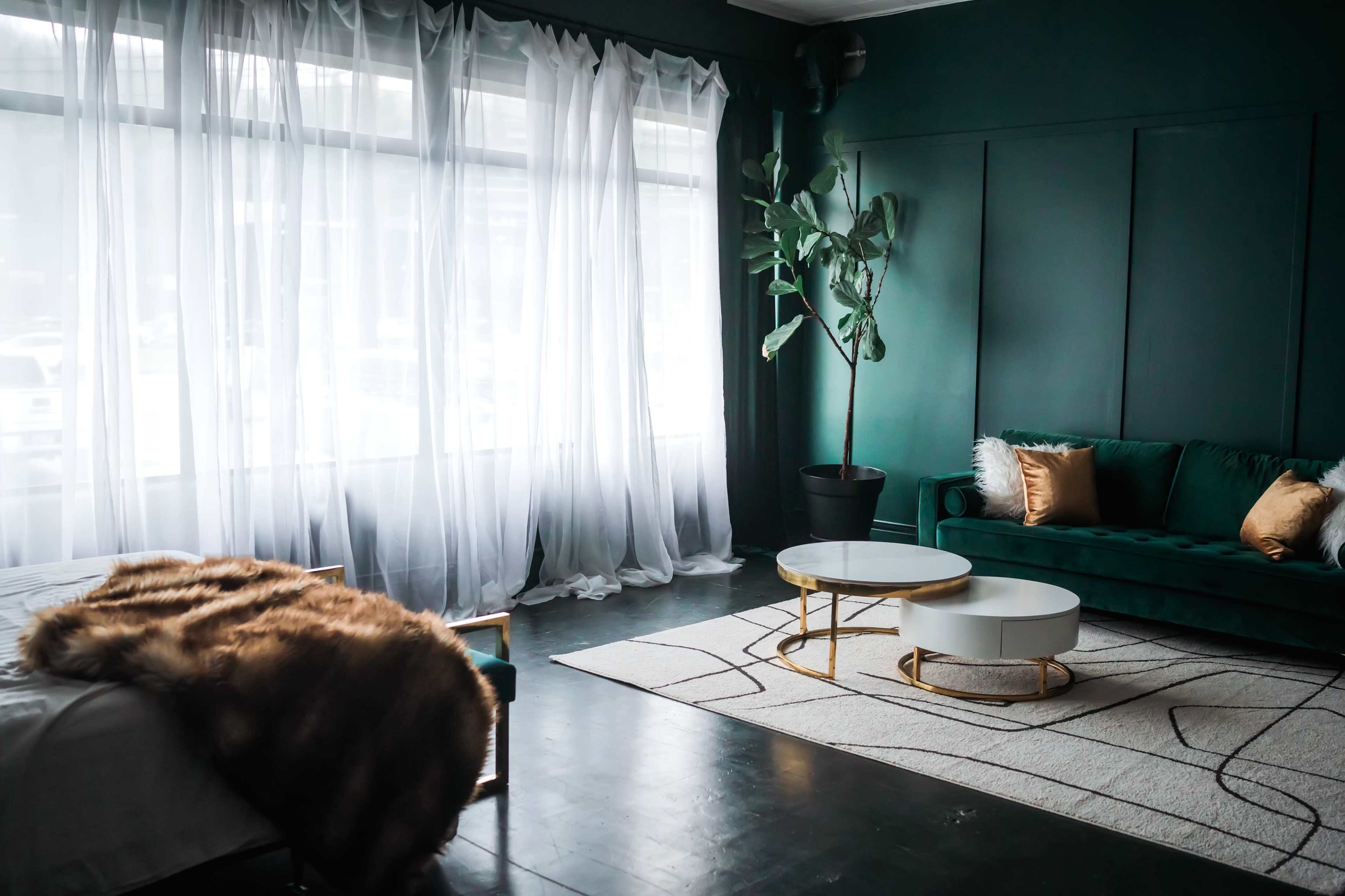 The image shows a stylish room featuring a green sofa, a round coffee table, and large windows covered with sheer white curtains.