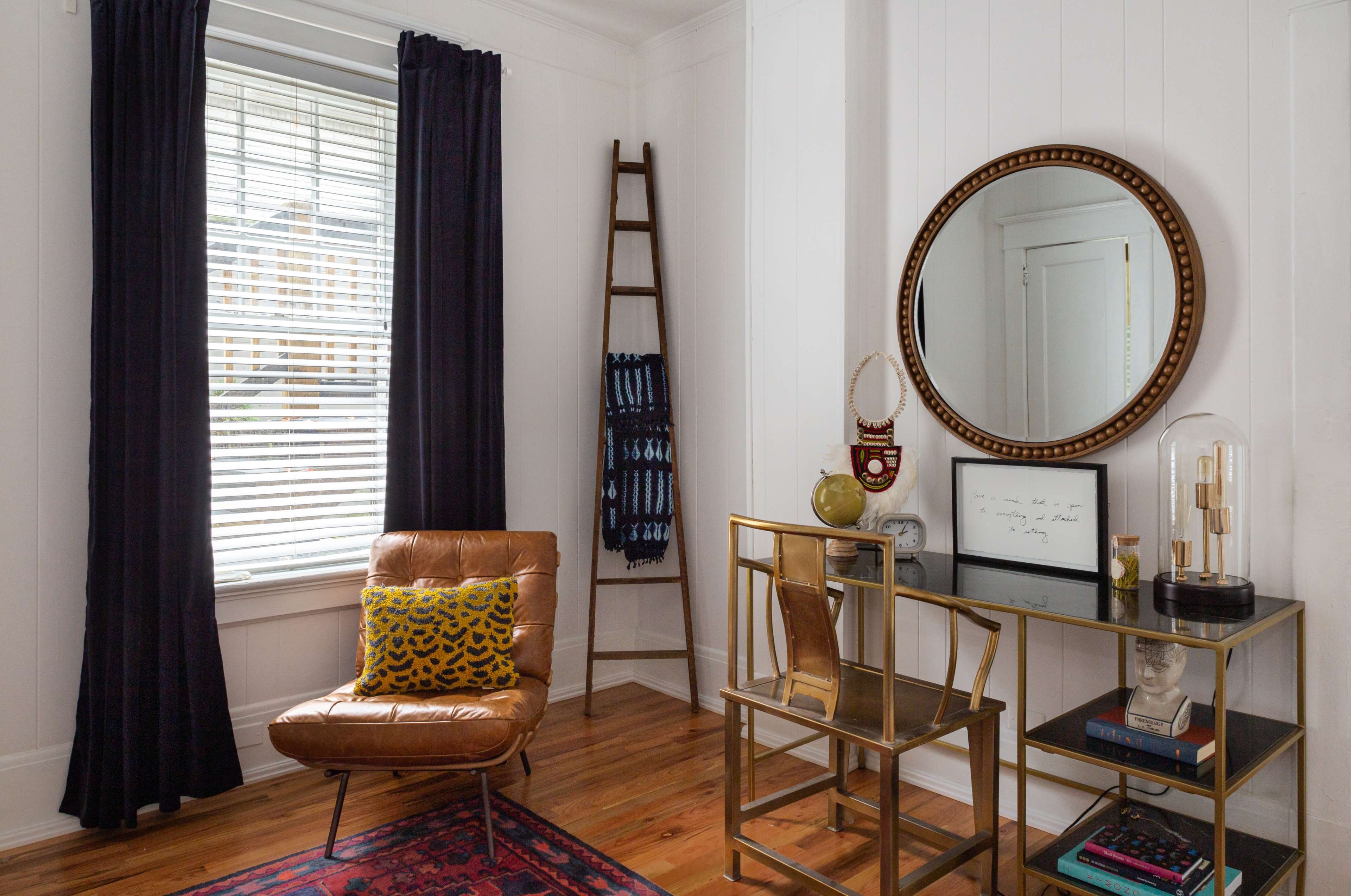 A cozy corner features a brown leather chair, a round mirror, a gold-framed console table with decorative items, and a wooden ladder leaning against the wall.