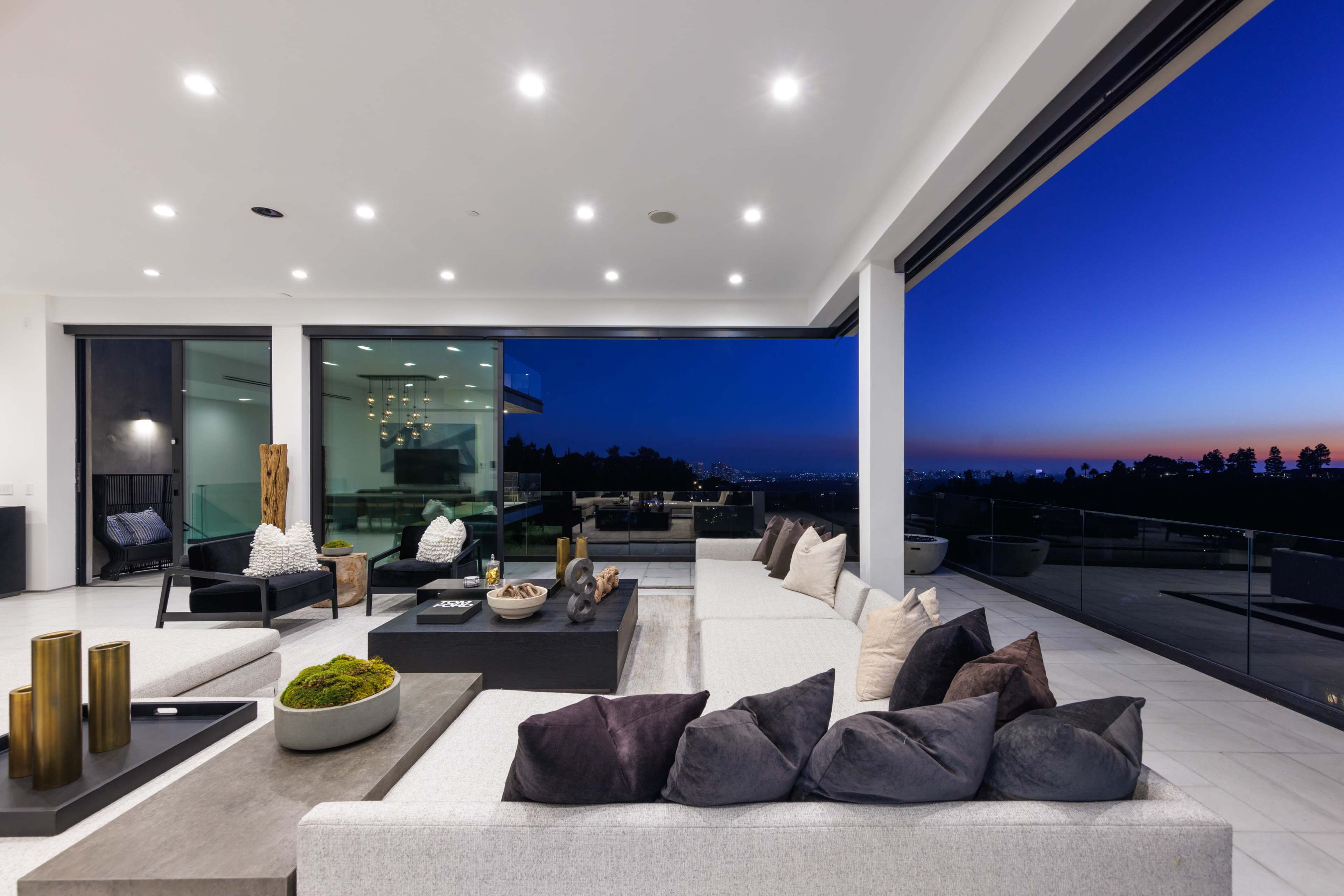 A modern living room features sleek furniture and large glass windows that open up to a panoramic view of a sunset cityscape.