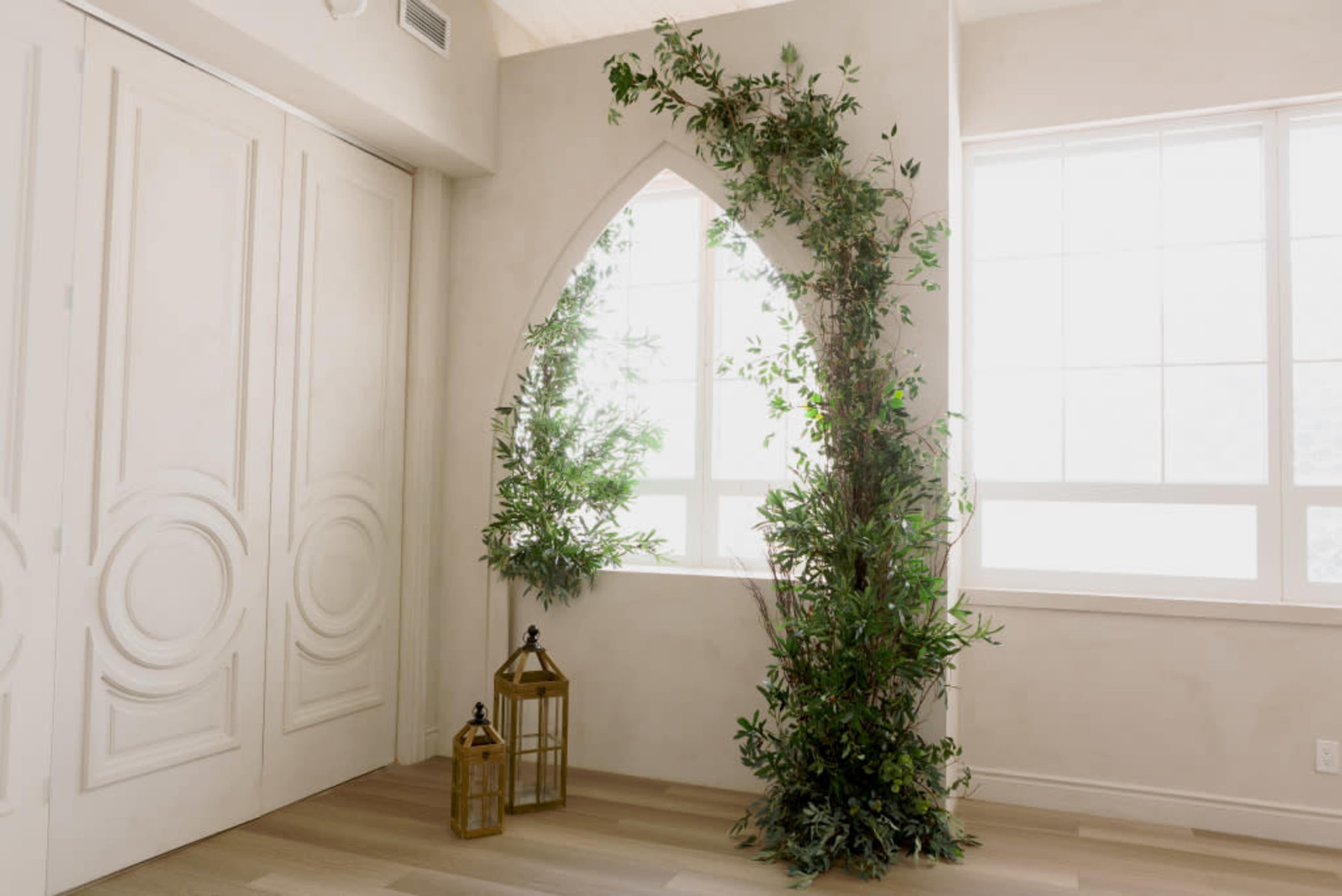 A light-filled room features an arched window surrounded by greenery and two decorative lanterns on the floor.