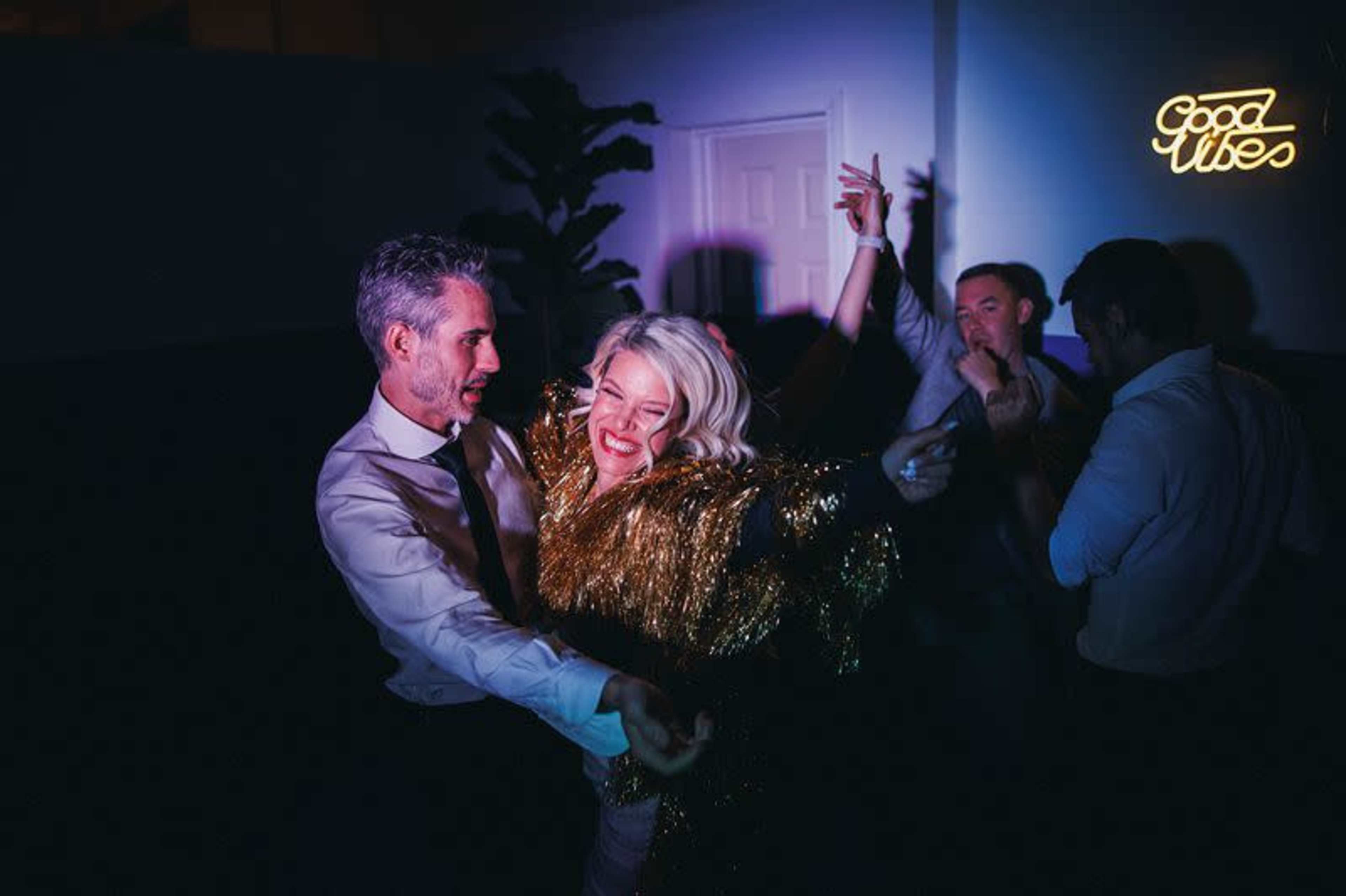 A group of four people dance energetically in a dimly lit room decorated with a "Good Vibes" sign.