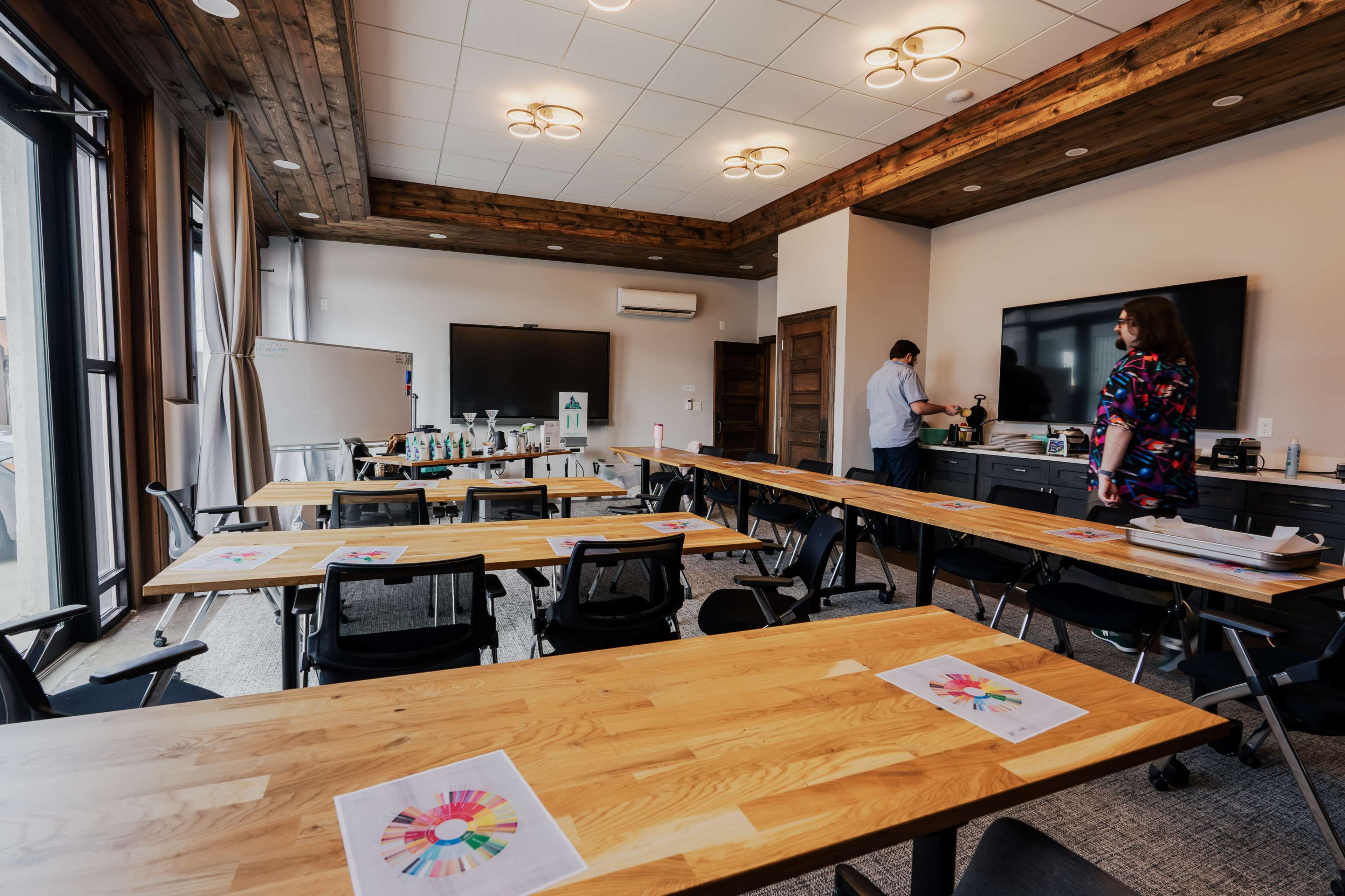 A modern meeting room features wooden tables arranged for a workshop, with colorful printed materials on each table and video equipment set up for presentations.