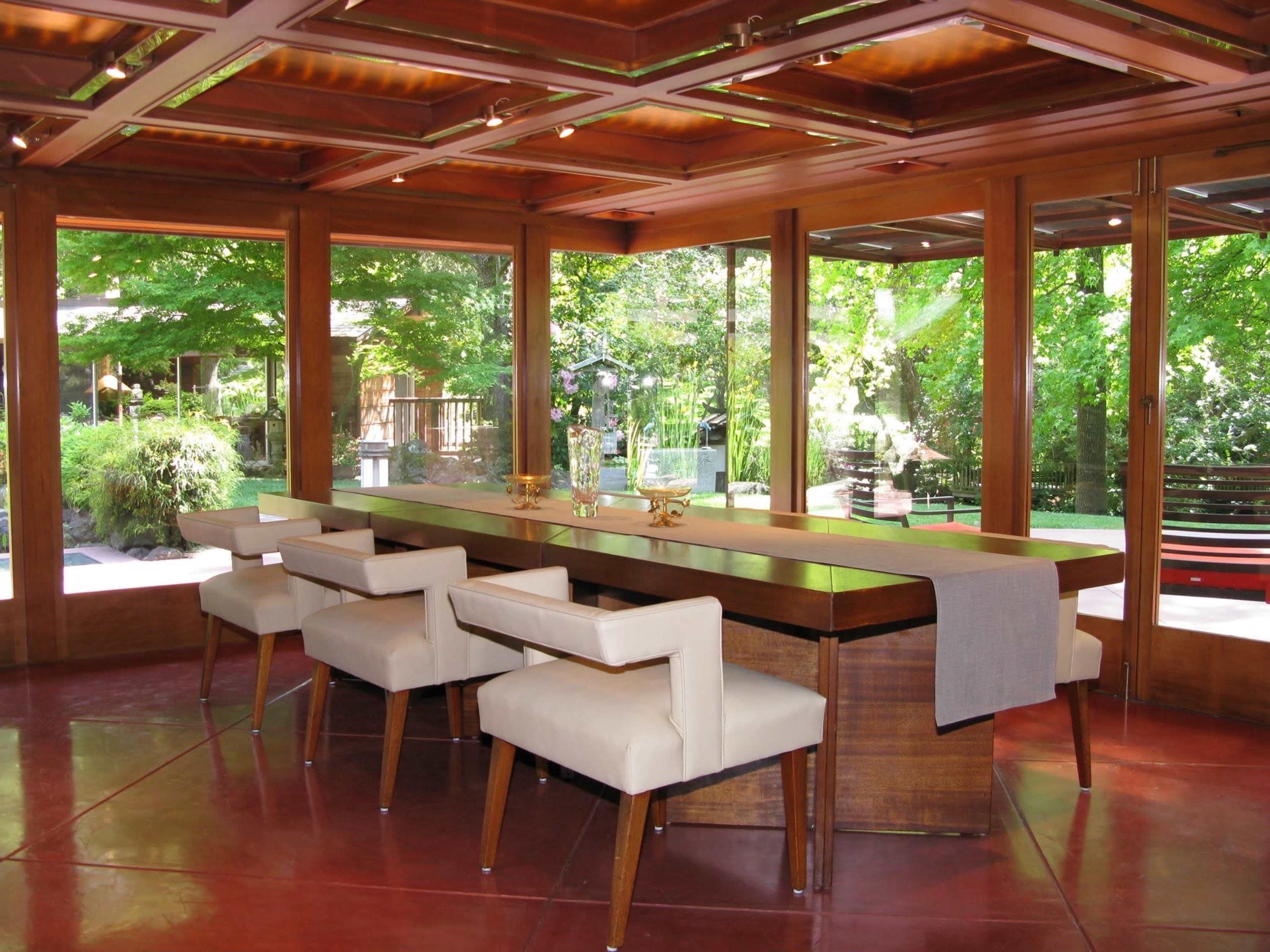 A contemporary dining area with a long wooden table surrounded by four upholstered chairs, overlooking a lush outdoor garden through large windows.