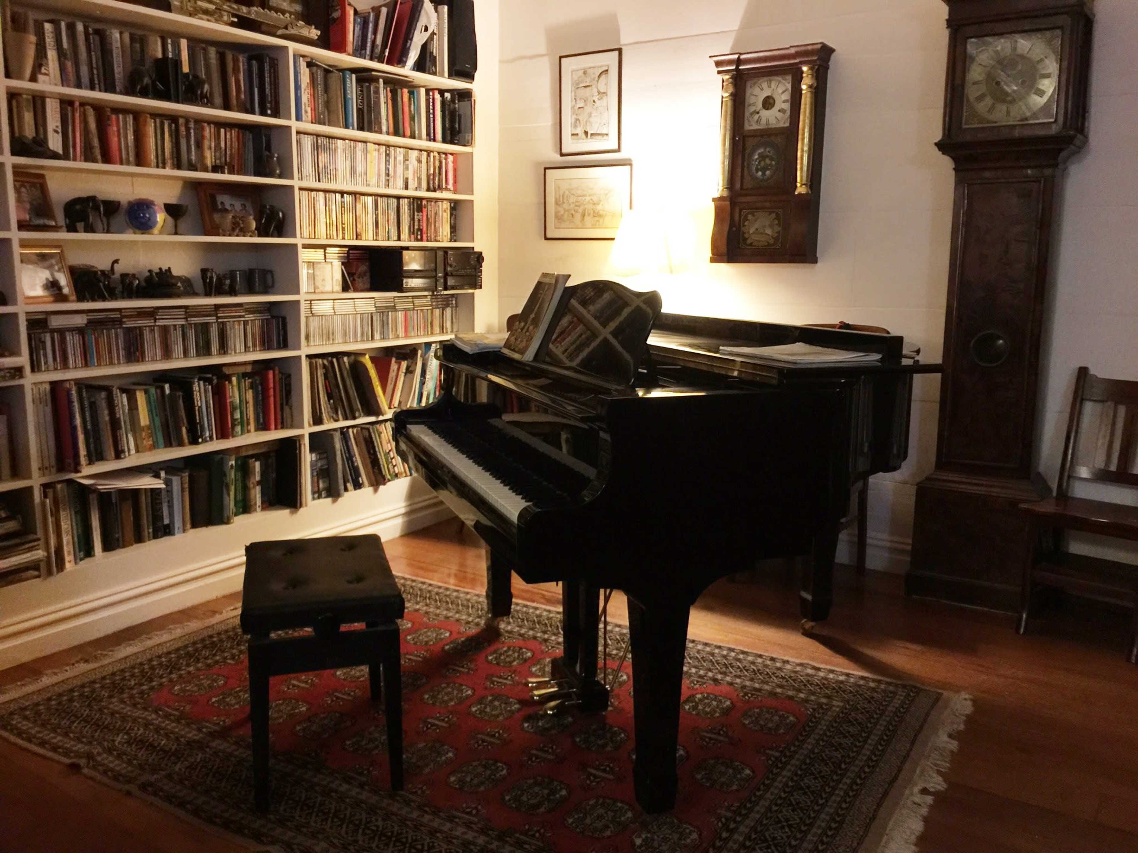 A grand piano sits next to a bookshelf filled with books, and a tall clock stands in the background of a cozy room.