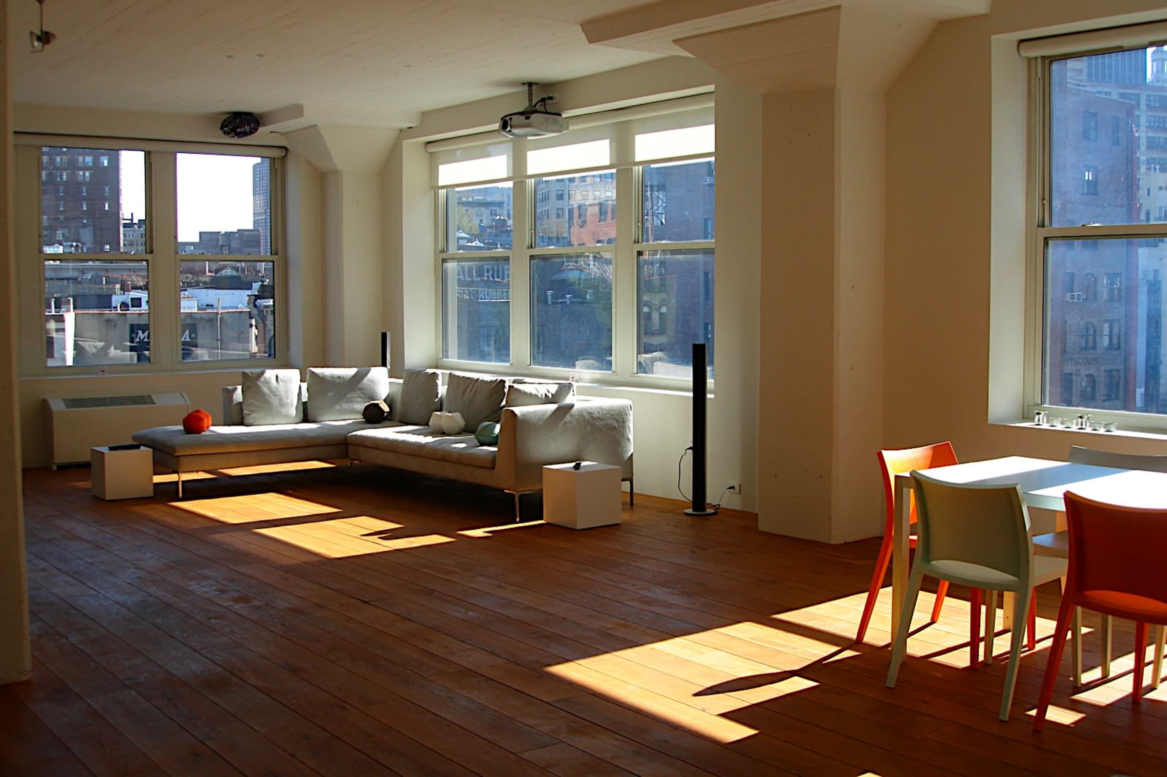 The image shows a spacious room with large windows allowing sunlight to illuminate a sectional sofa, a small table, and a dining area with chairs on a wooden floor.