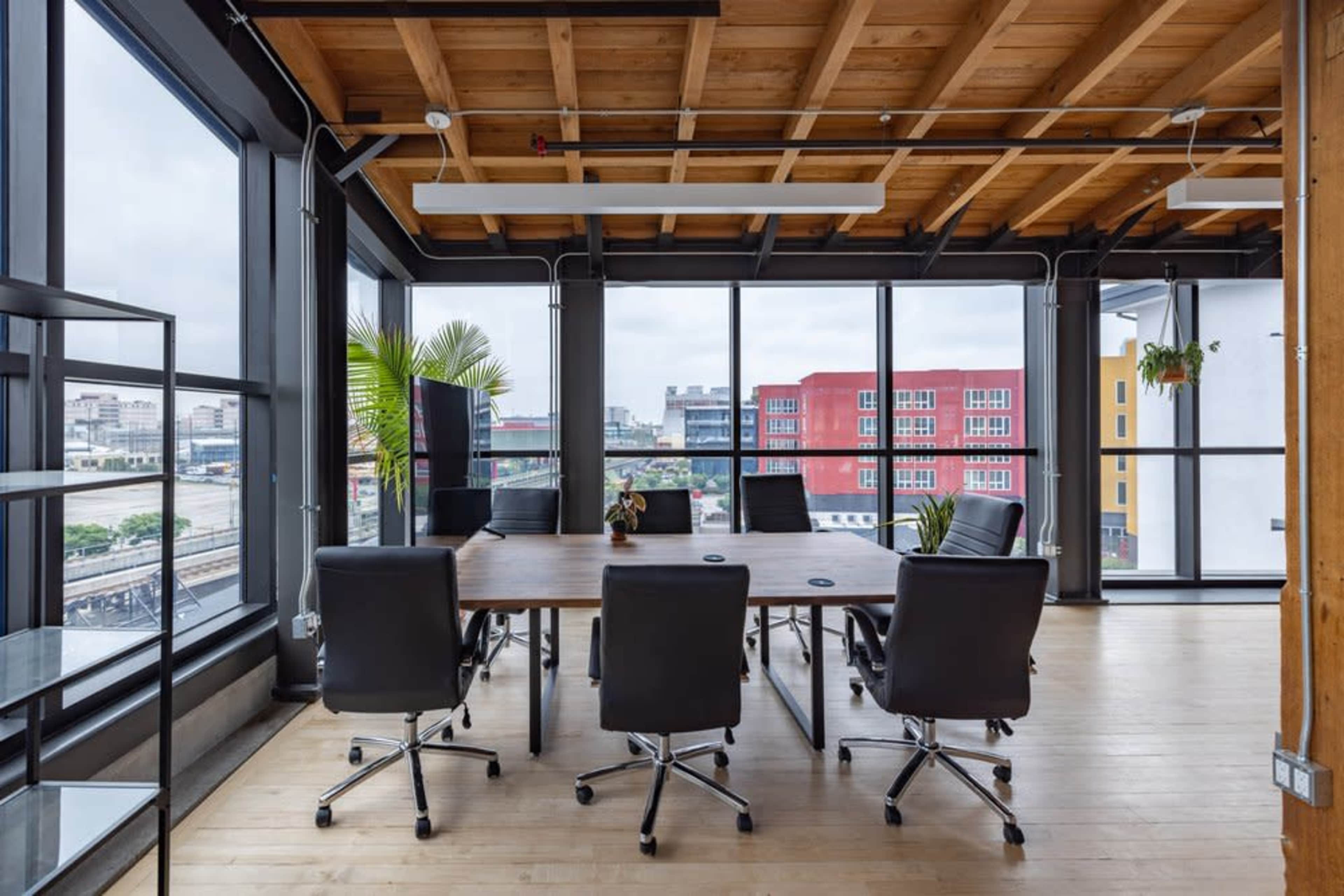 The image shows a modern conference room with a large wooden table surrounded by black chairs, featuring large windows that overlook an urban landscape.