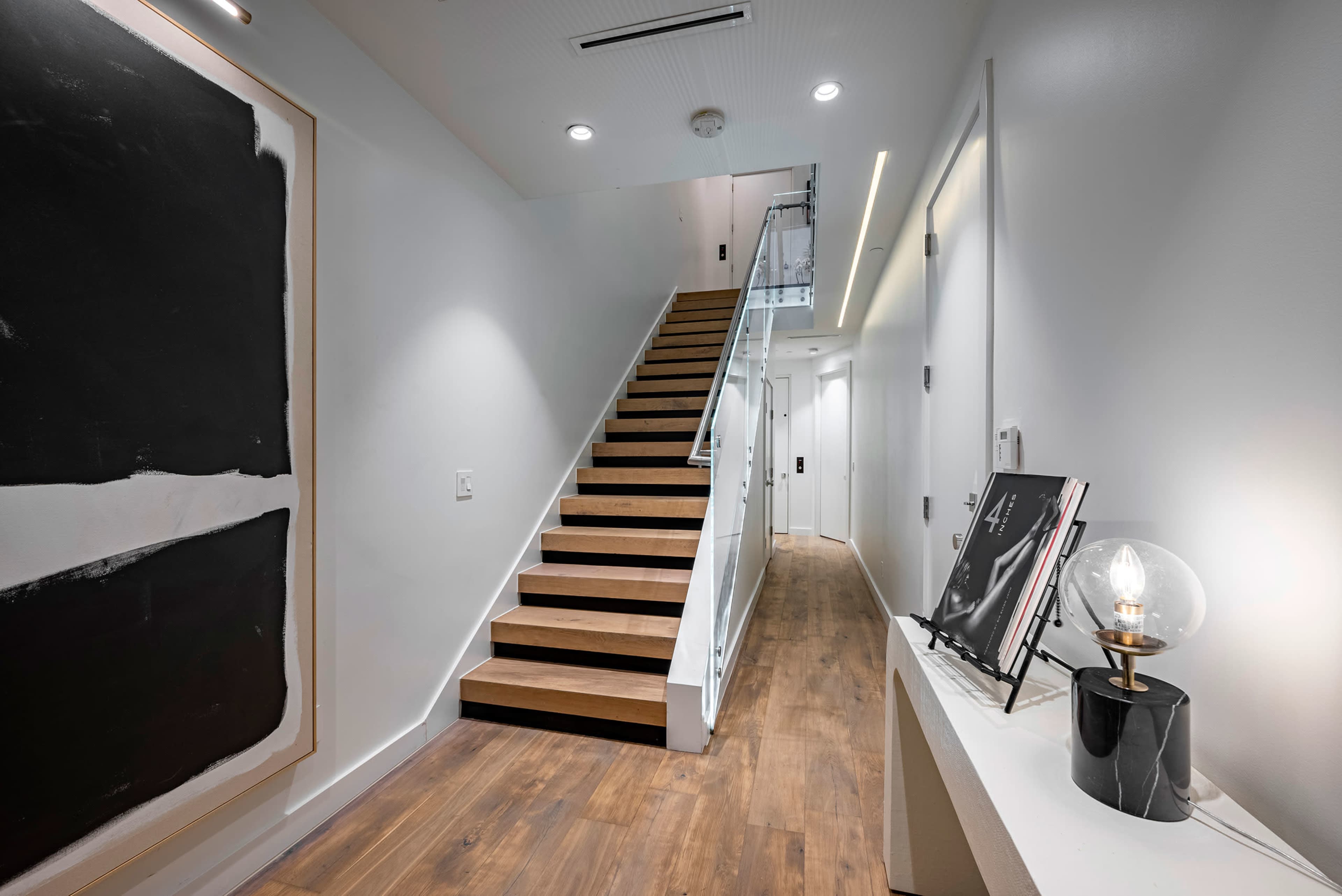 A modern hallway featuring a staircase with wooden steps, a large black and white artwork, and a small table with a lamp and a picture frame.