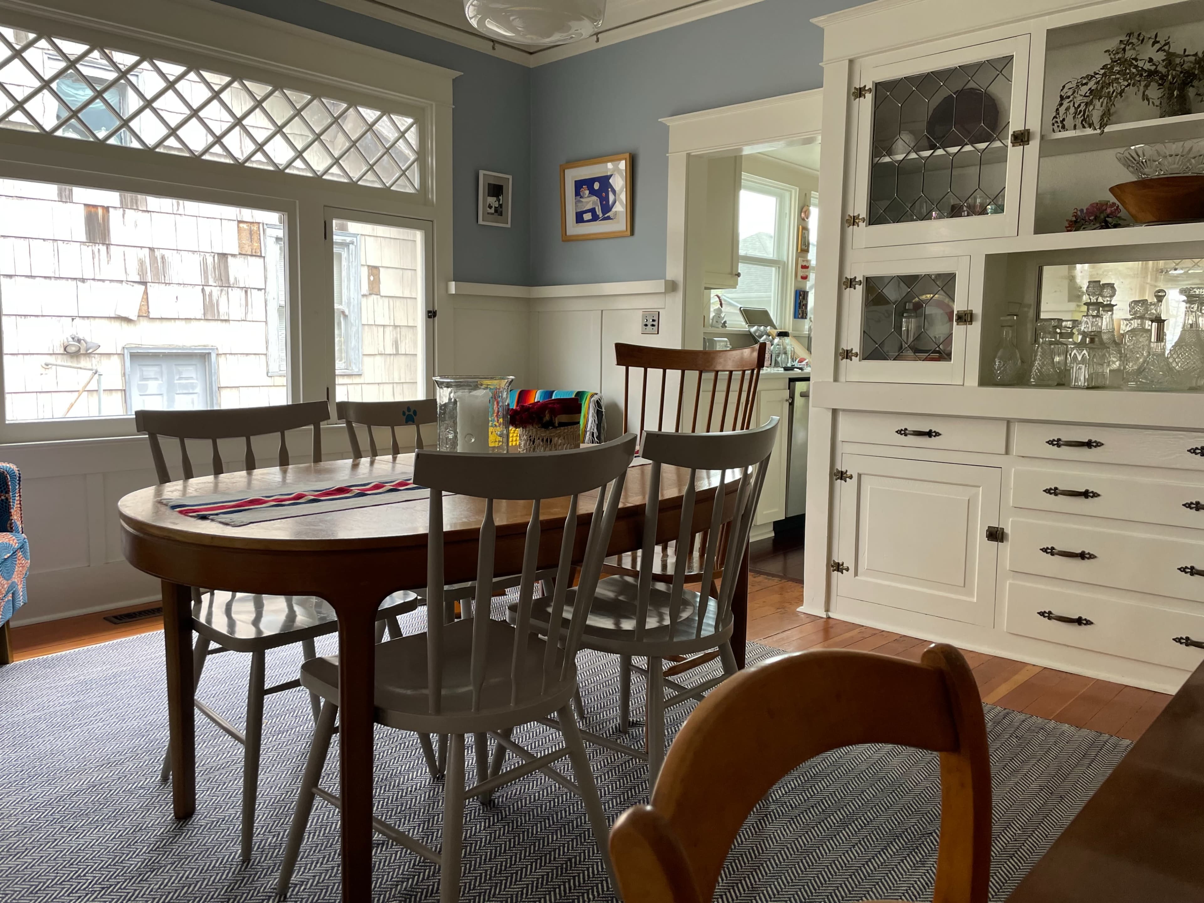 A dining room features a wooden table surrounded by six chairs, with a buffet cabinet and large windows allowing natural light.