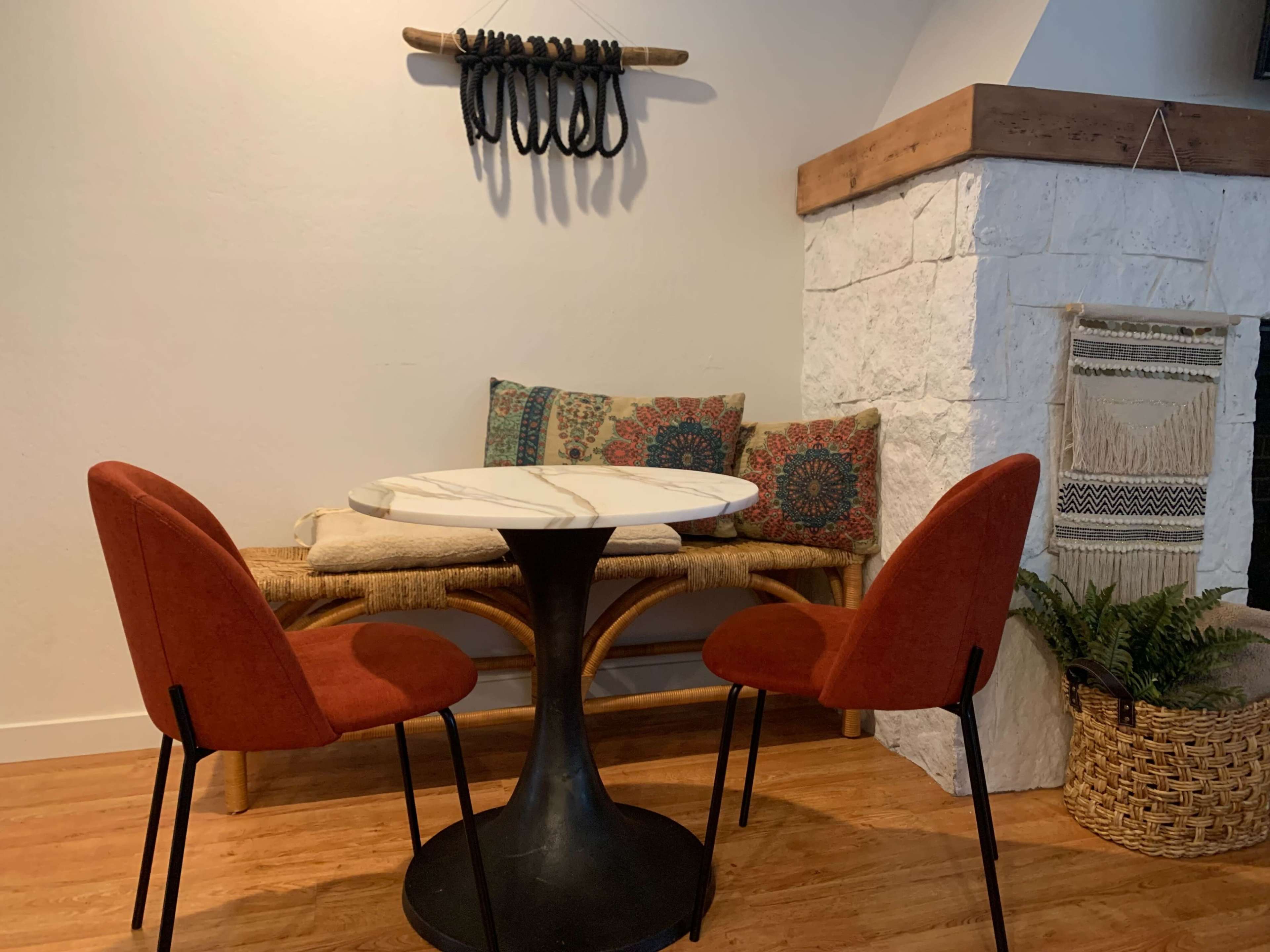 A small dining area features a round marble table with two red chairs, a bench with patterned cushions, and a stone fireplace in the background.