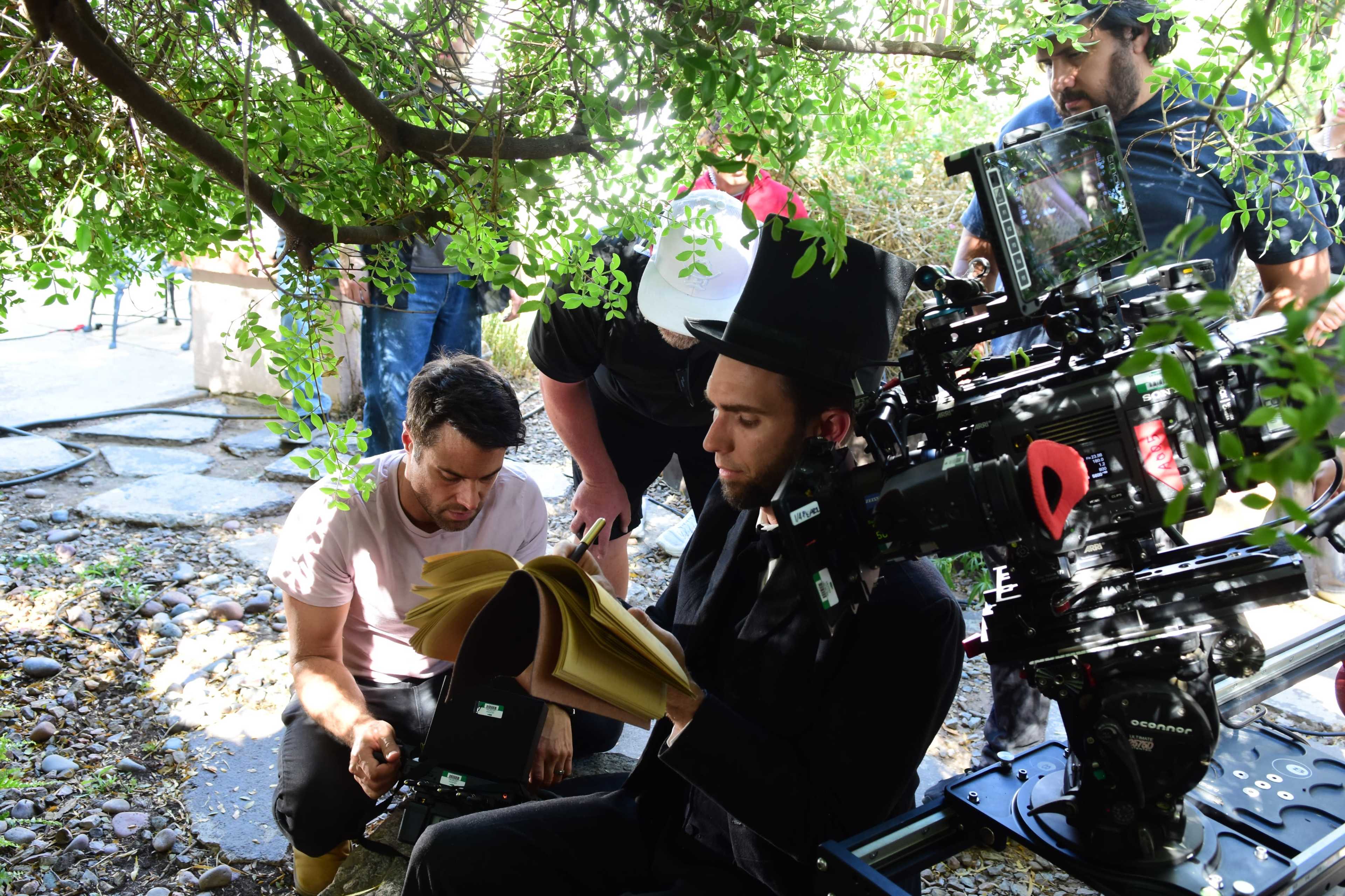 A film crew is working under a tree, with one person reading a script while another adjusts camera equipment.