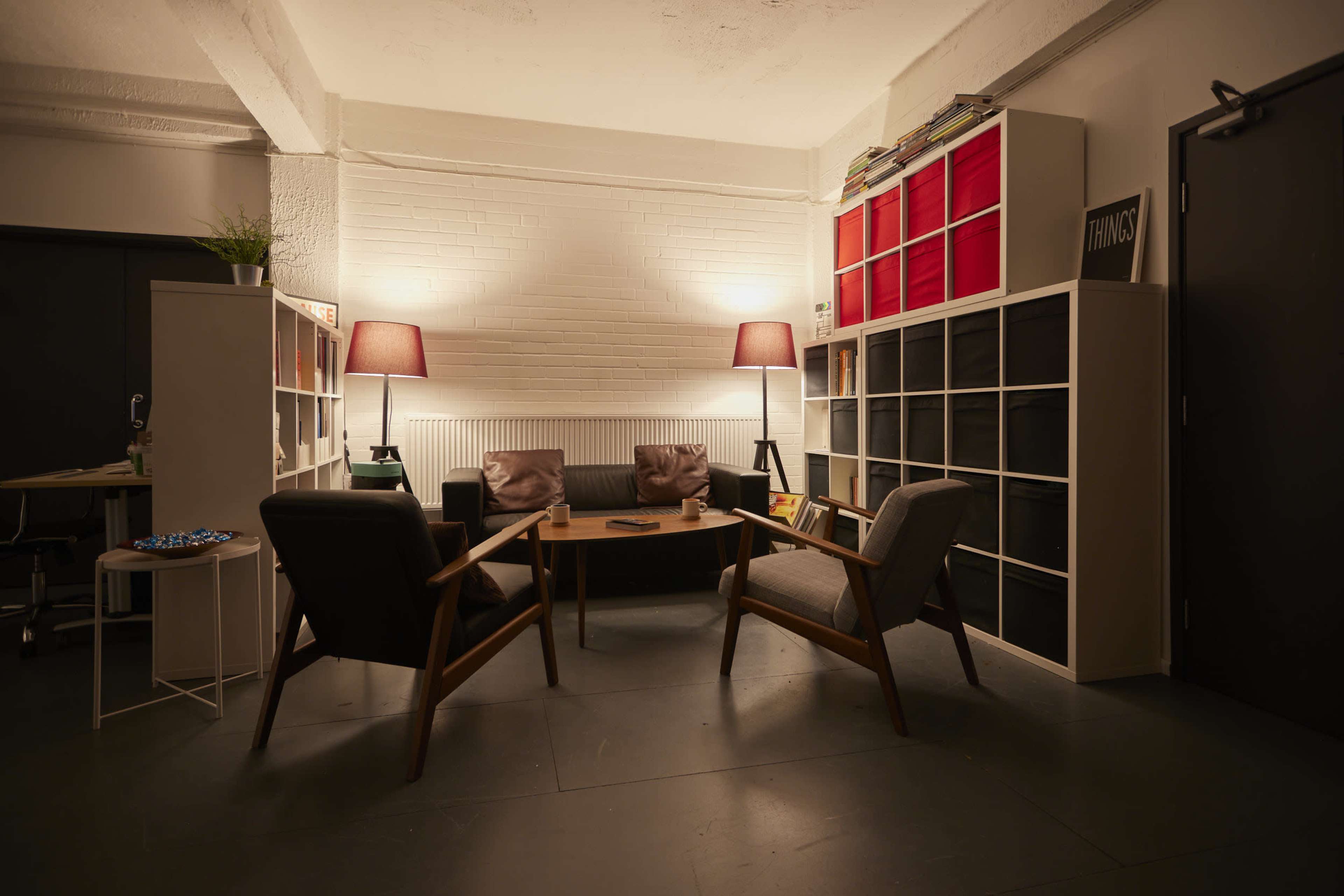 A cozy office space features a table surrounded by two armchairs and two lamps, with shelving units and a desk in the background.