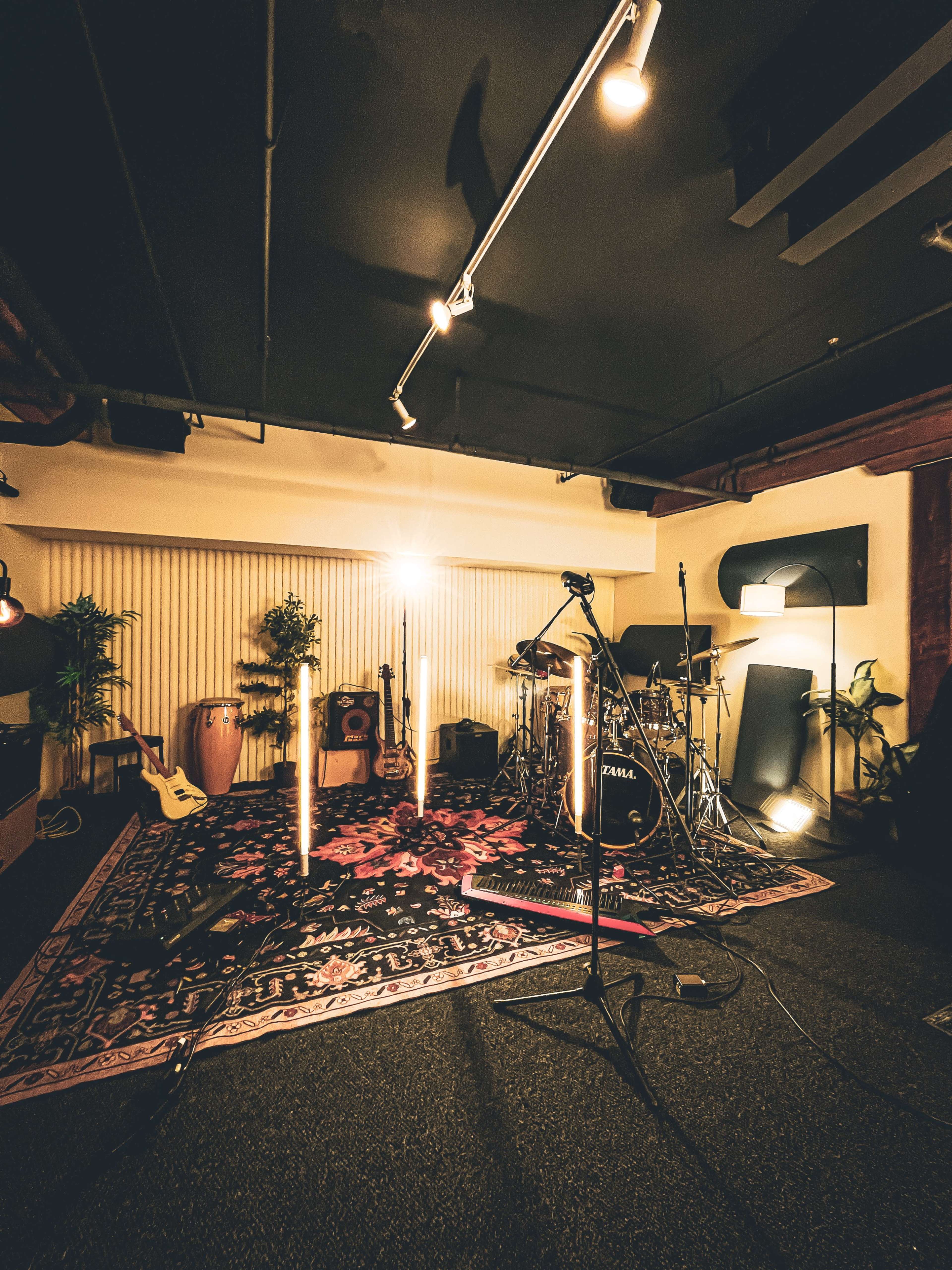 The image shows a music rehearsal space featuring various instruments, including a drum set and guitars, arranged around a decorative rug under warm lighting.