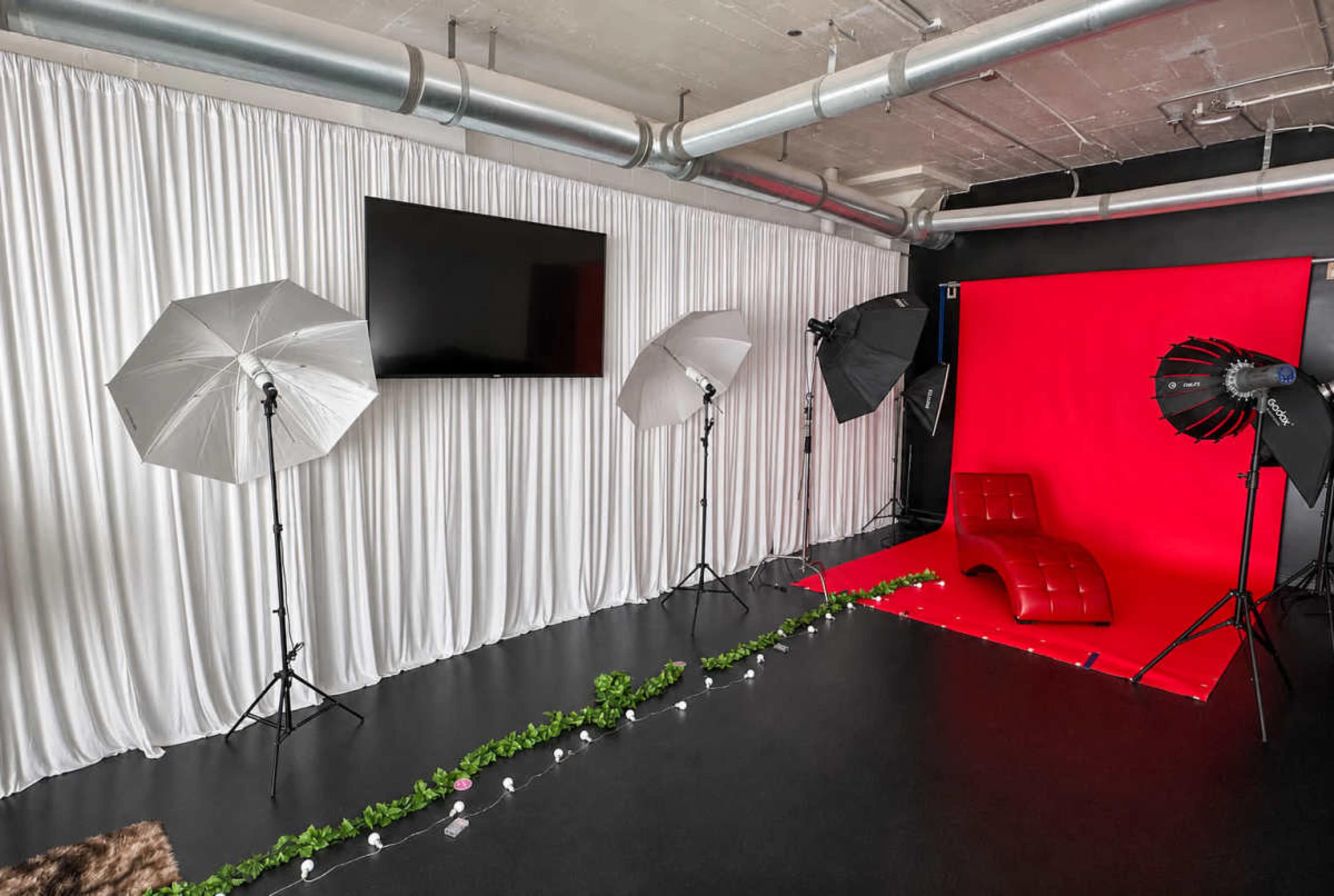 The image shows a photography studio equipped with various lighting equipment, a red backdrop, and a curved red sofa.