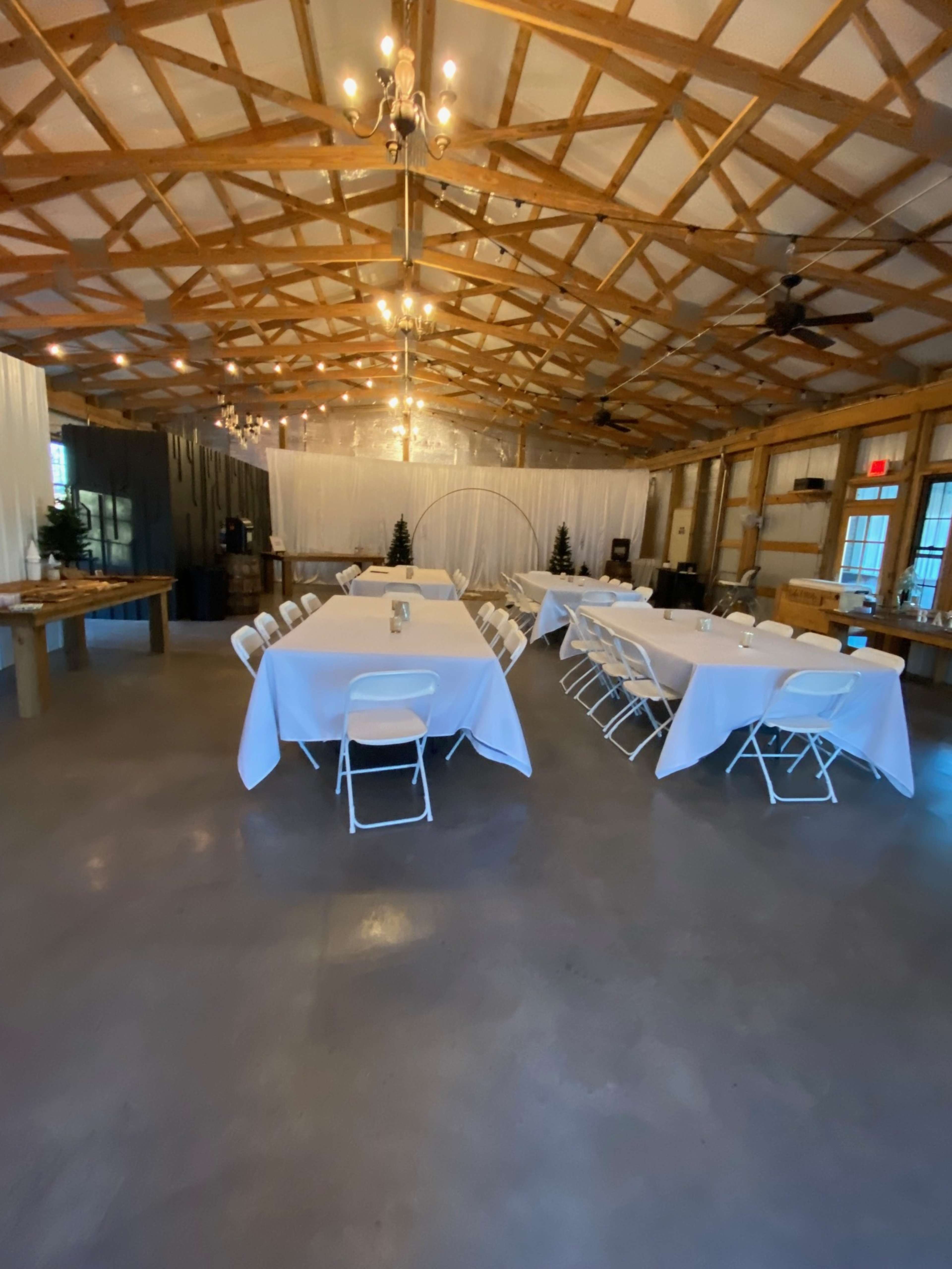 A spacious indoor event venue with several tables set up in a U-shape and string lights hanging from a wooden ceiling.