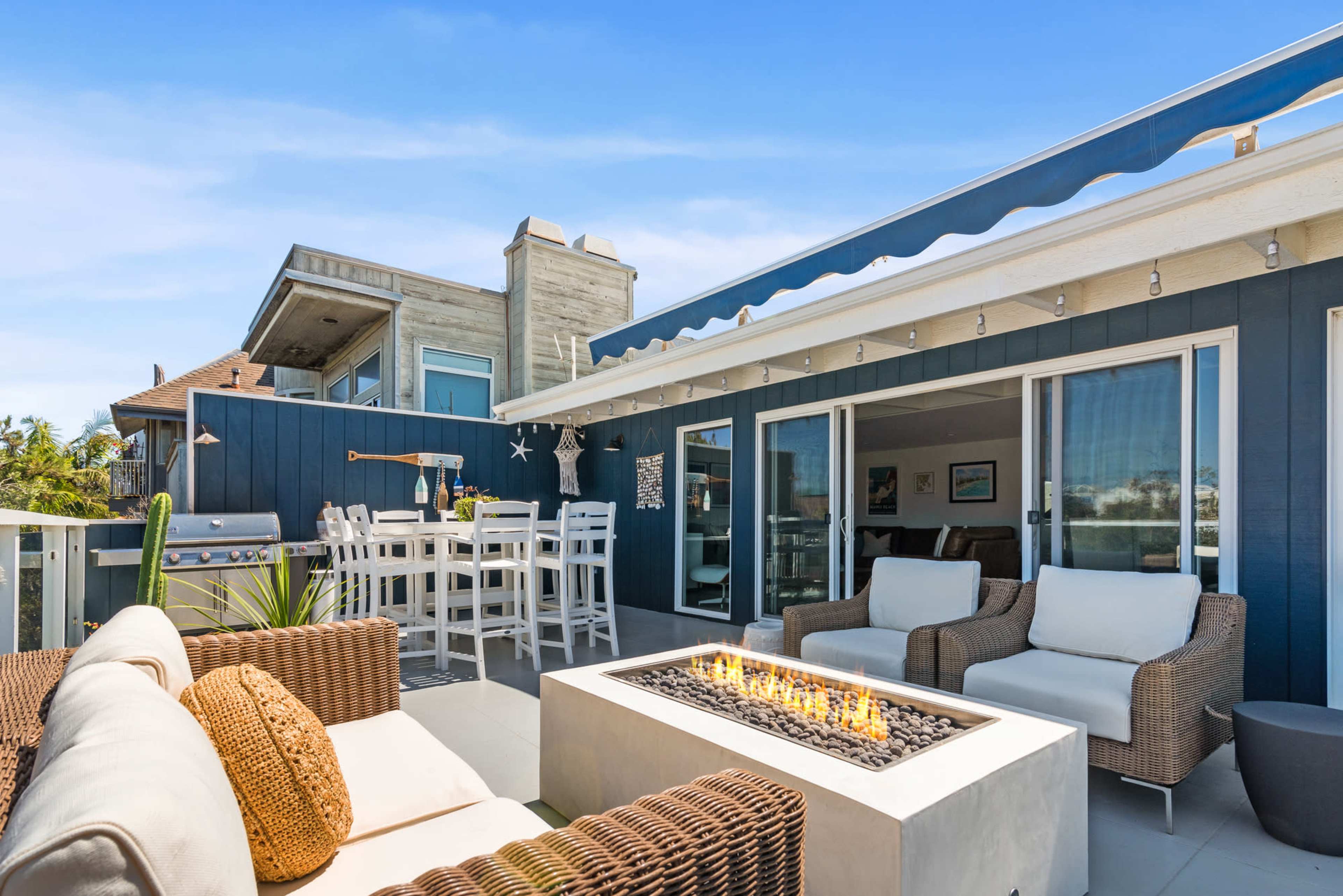 A modern outdoor patio features a fire pit surrounded by wicker seating, a dining area, and a barbecue grill, set against a backdrop of contemporary homes.