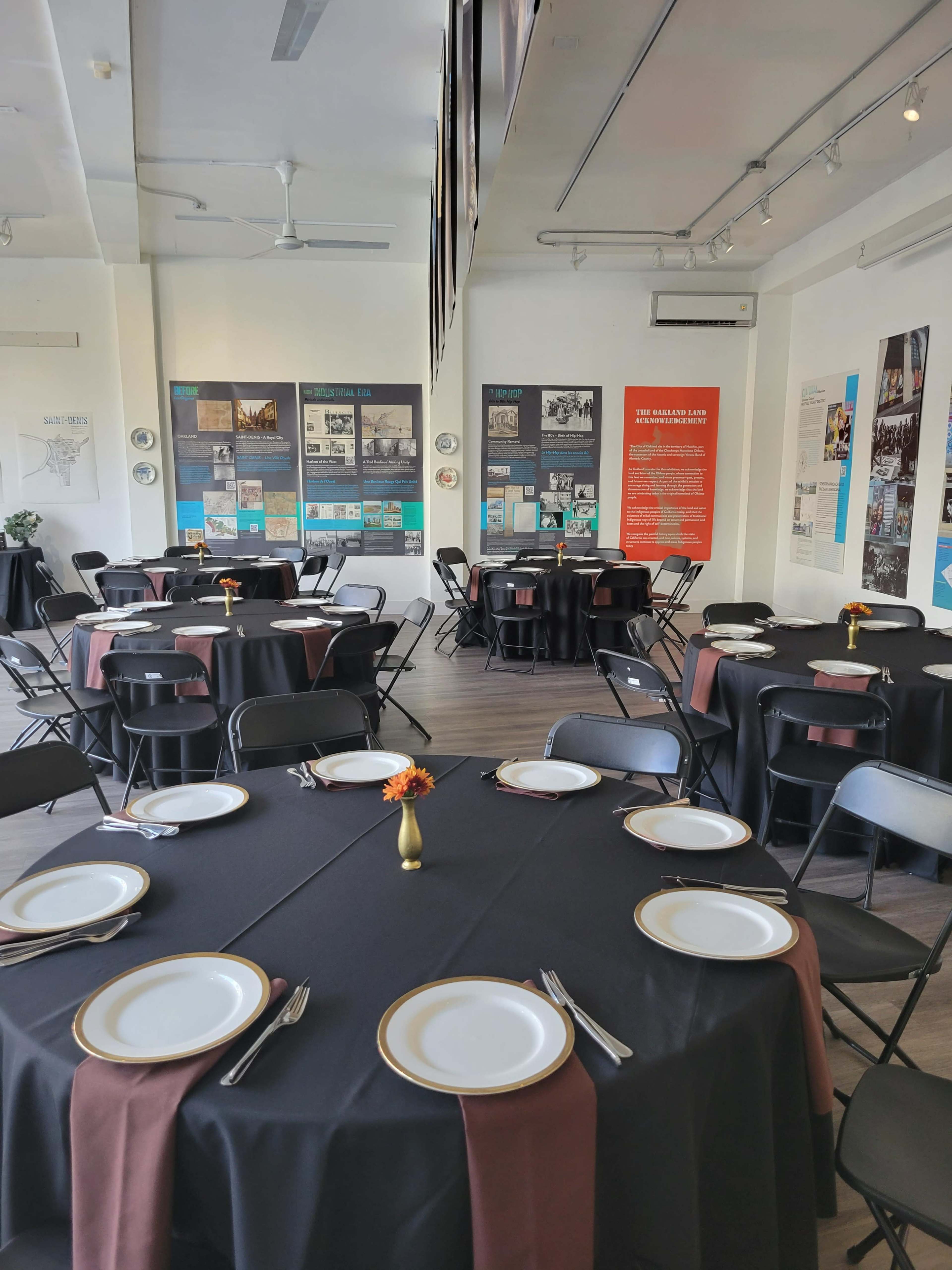 A dining area arranged with round tables covered in black tablecloths, each set with white plates, silverware, and a small decorative vase at the center.