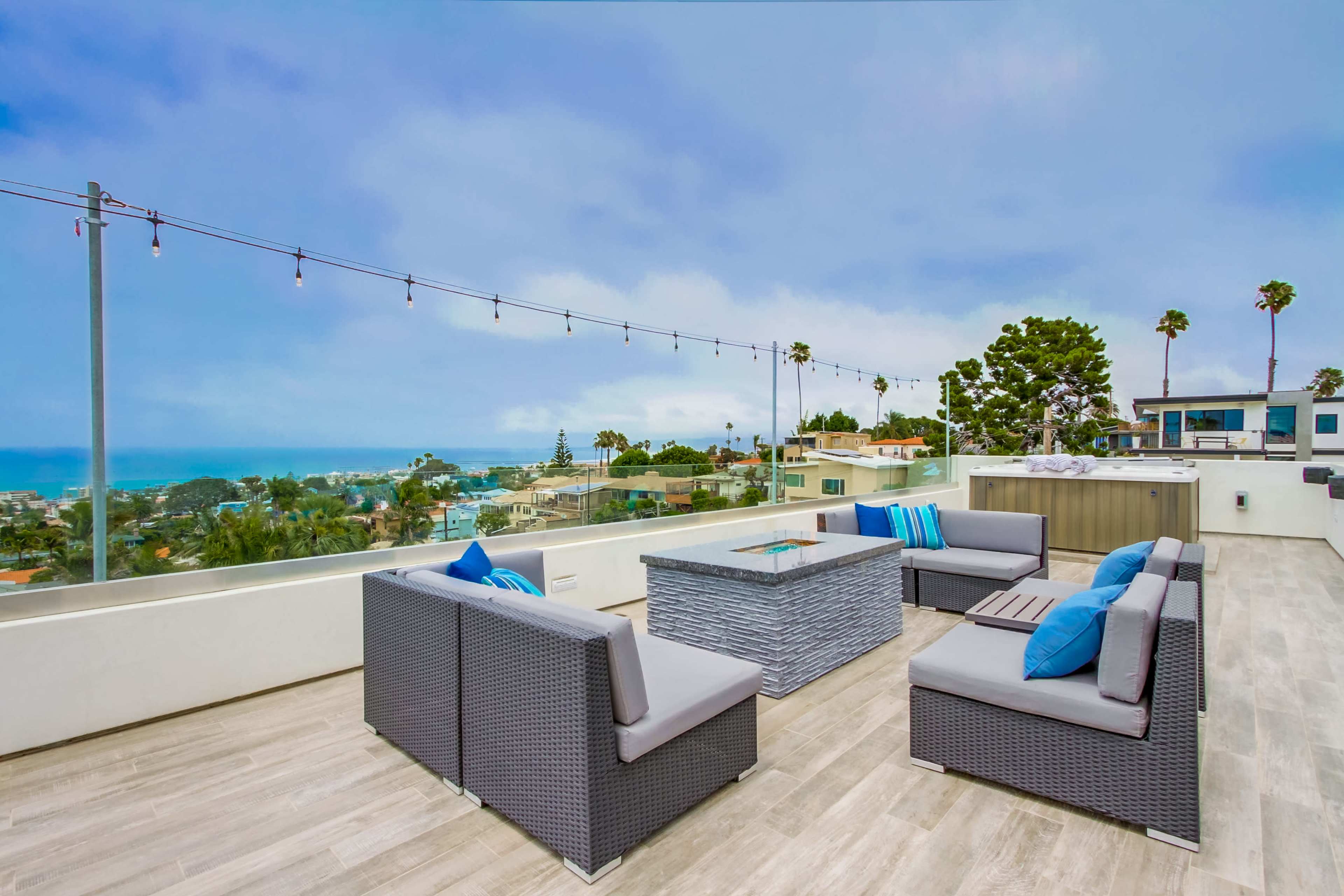 A rooftop patio features gray seating with blue cushions, a central fire pit, and views of a coastal landscape under an overcast sky.