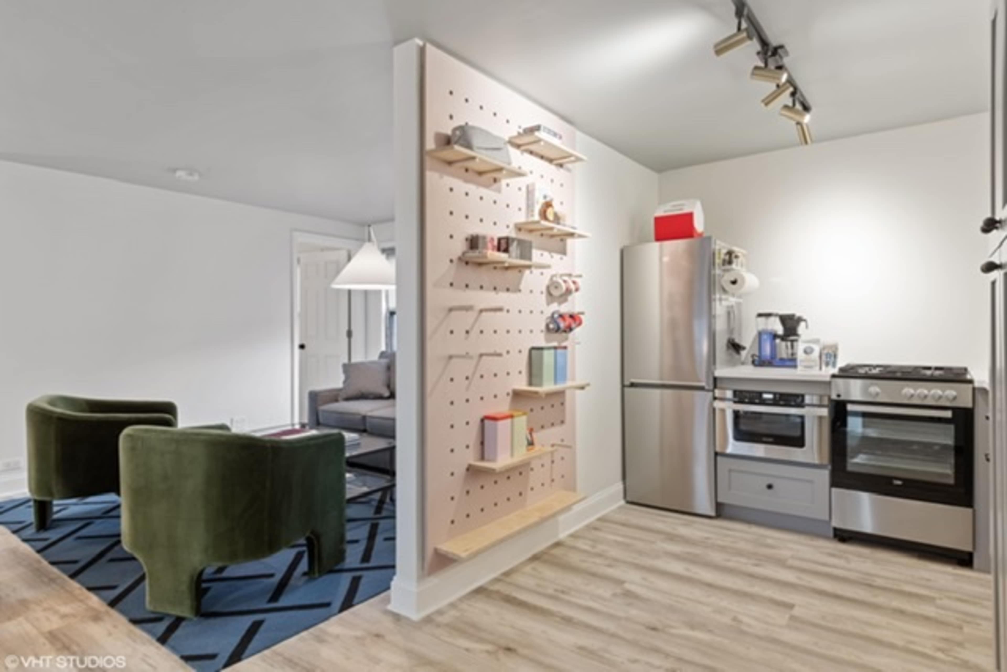 The image shows a modern kitchen and living area featuring a pegboard shelf, stainless steel appliances, and two green armchairs on a patterned rug.