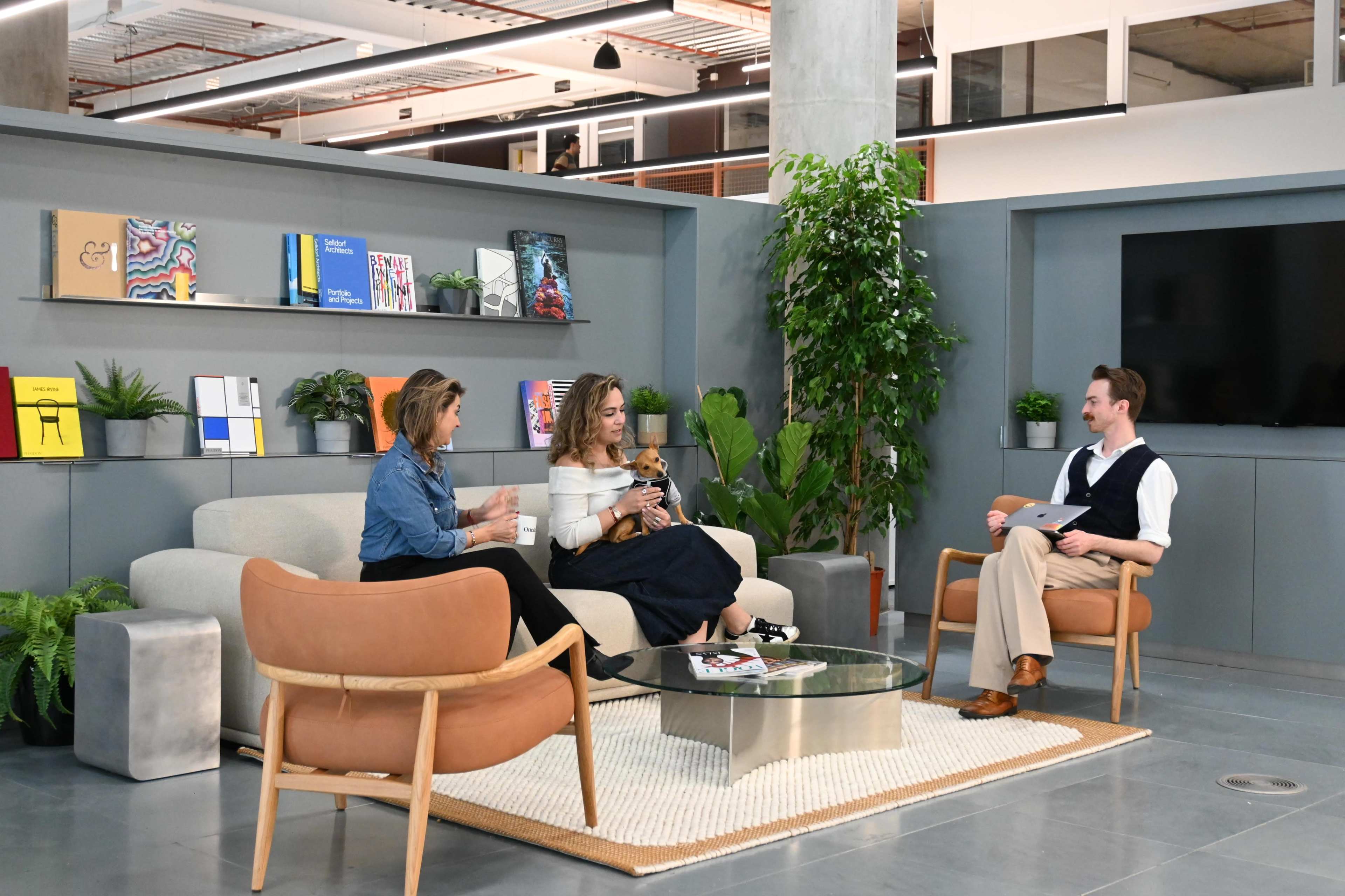 Three people are seated in a modern lounge area, engaged in conversation, with plants and books on display in the background.