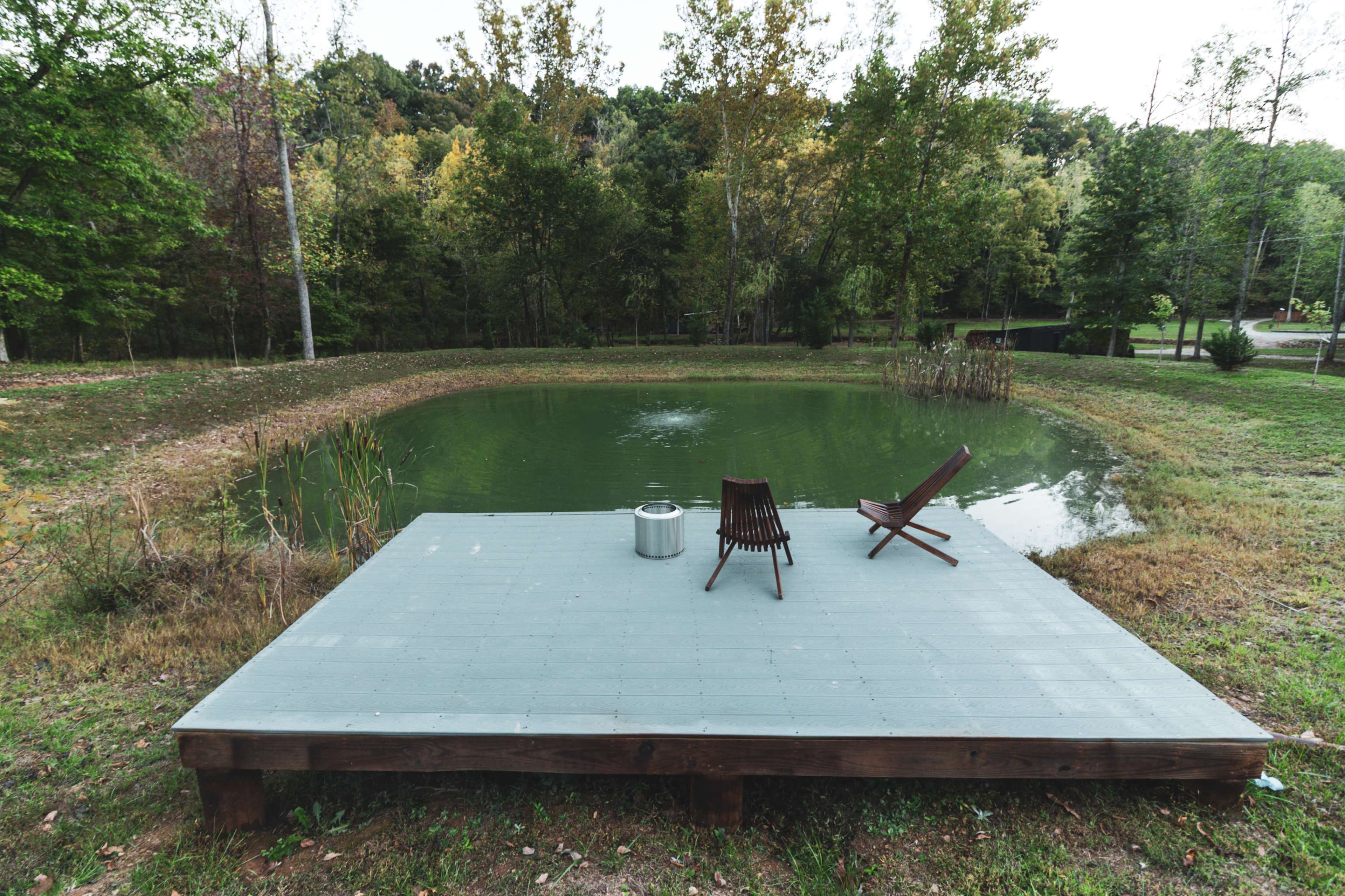 A wooden deck with two chairs overlooks a green pond surrounded by trees.