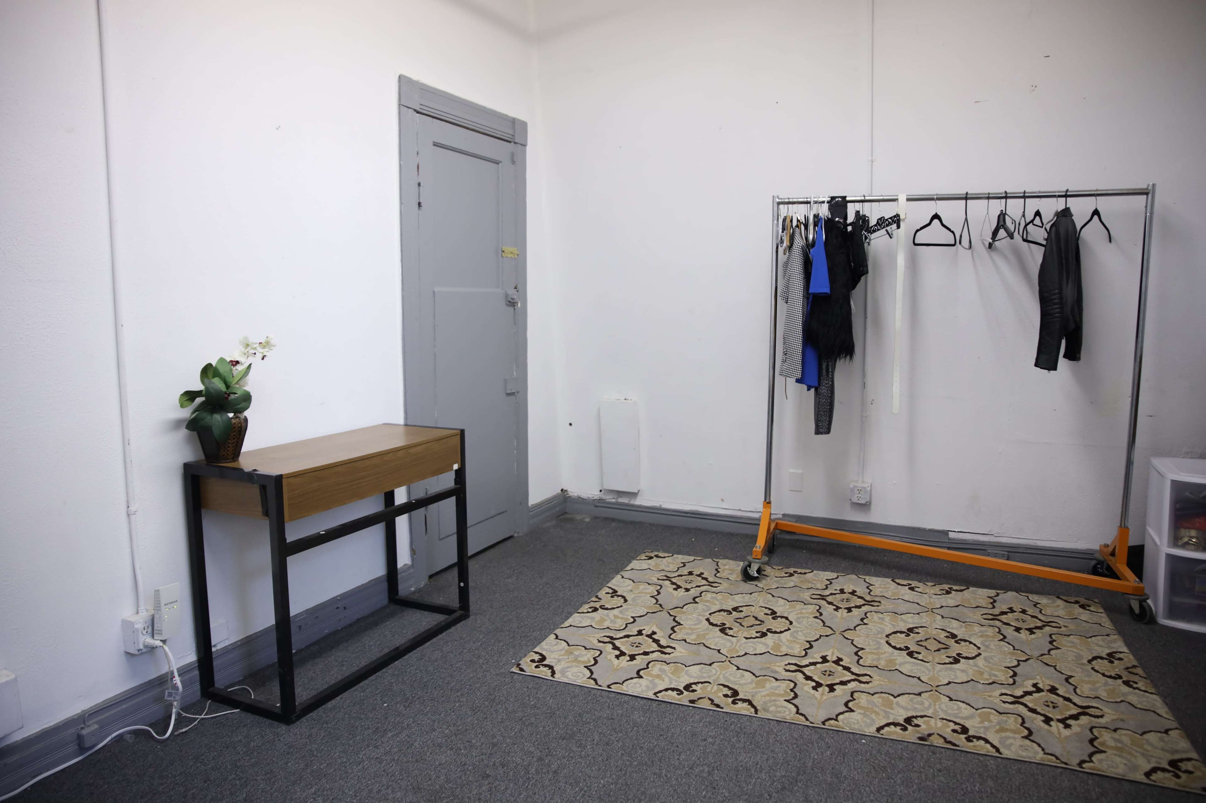 A minimalist room with a small wooden table, a clothing rack holding several garments, a patterned rug, and a door.
