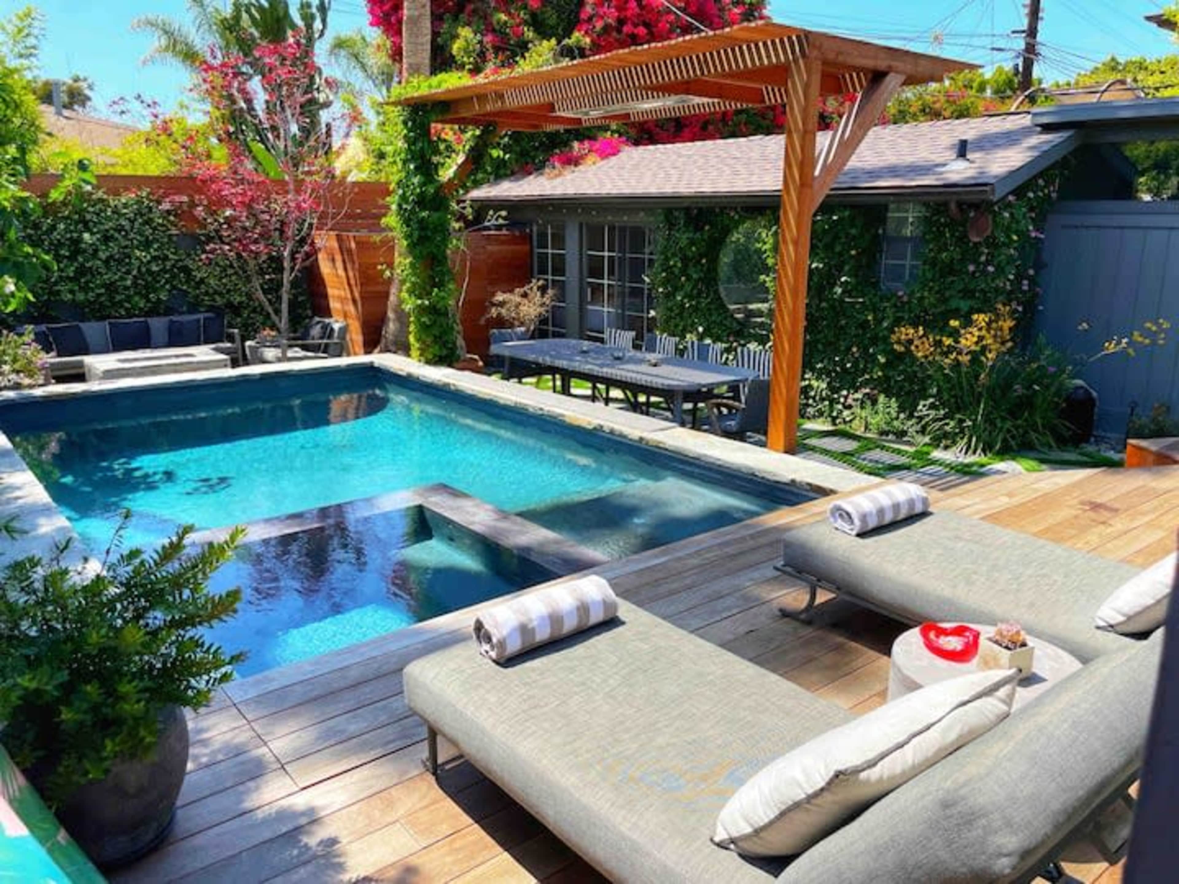 The image shows a modern backyard pool area featuring a rectangular pool surrounded by wooden decking, loungers, and greenery.