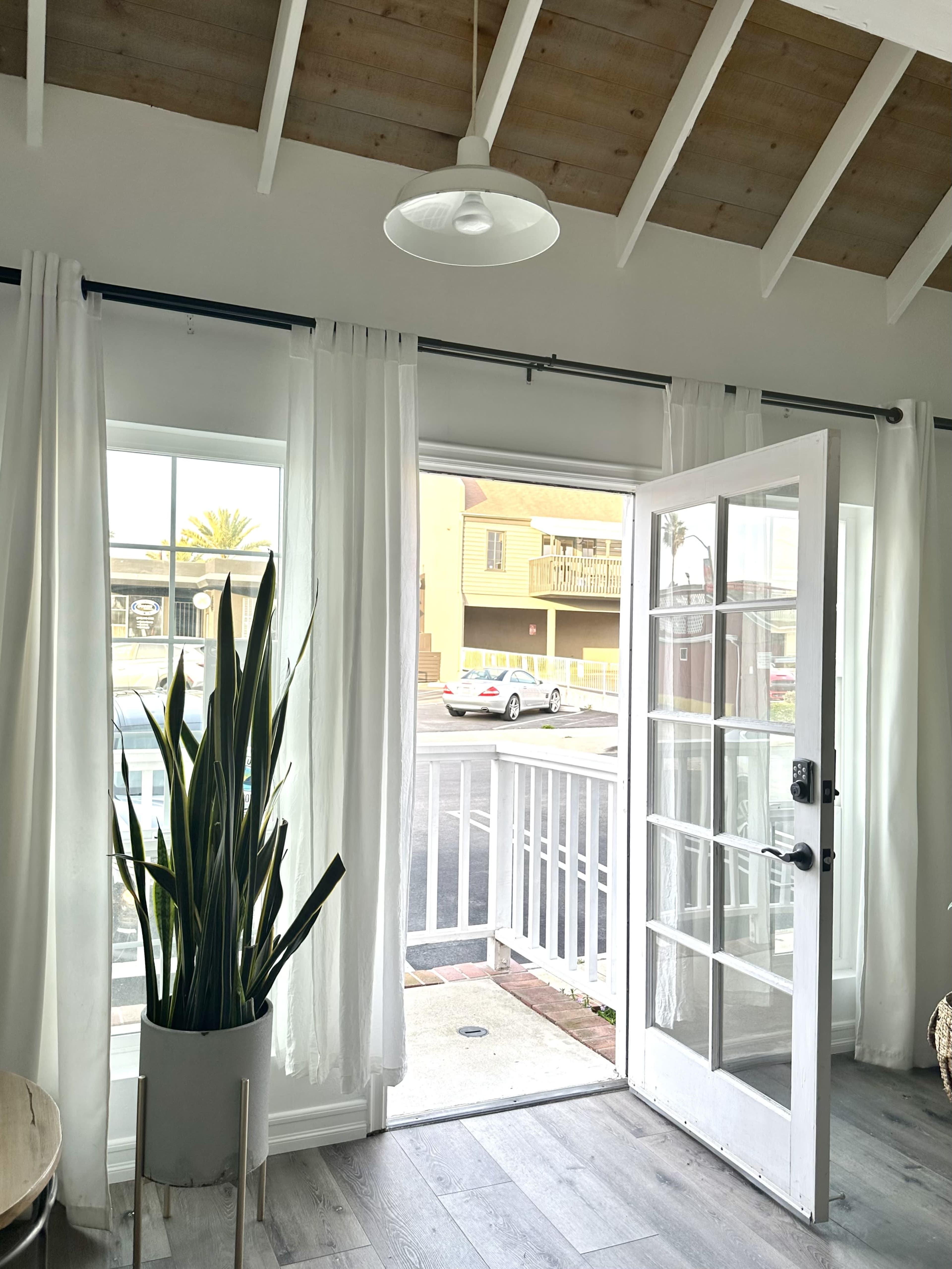 A bright interior features white curtains framing an open doorway that leads to an outdoor area with a parked car visible.