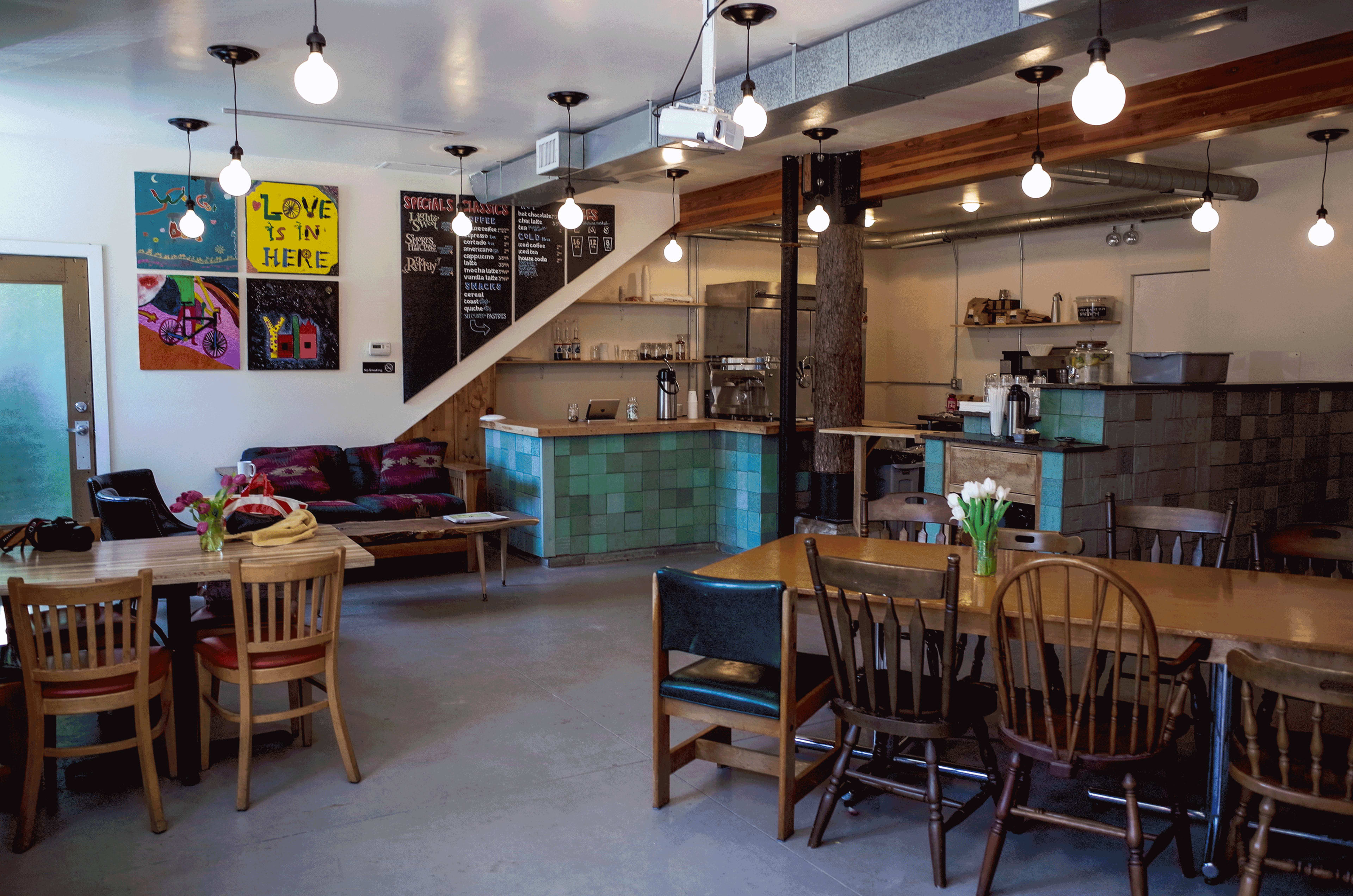 A cozy café interior with wooden tables, colorful chairs, a counter with a menu board, and decorative artwork on the walls.